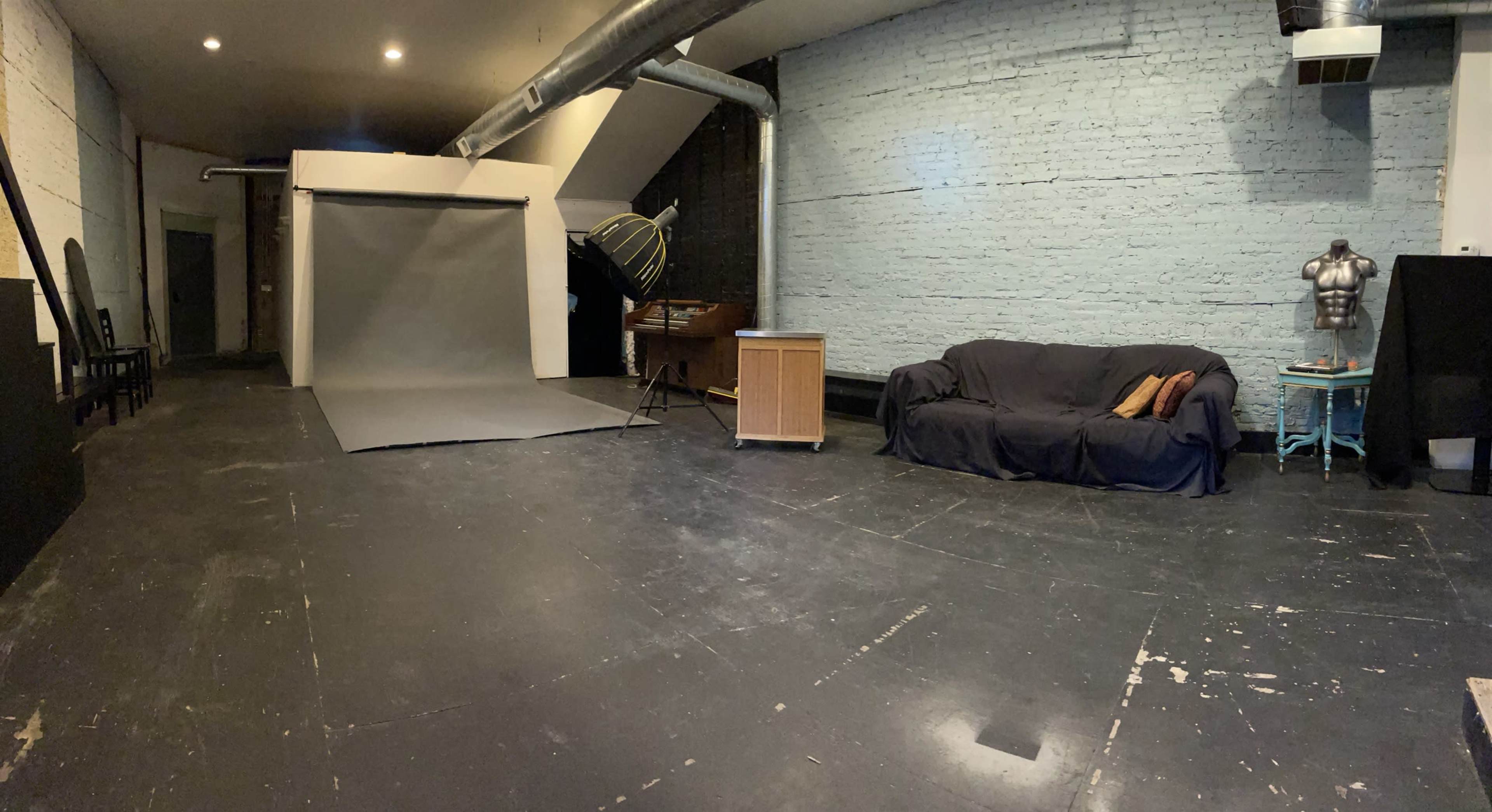 The image shows an empty studio space featuring a backdrop setup, a wooden cabinet, a black couch, and exposed brick walls.