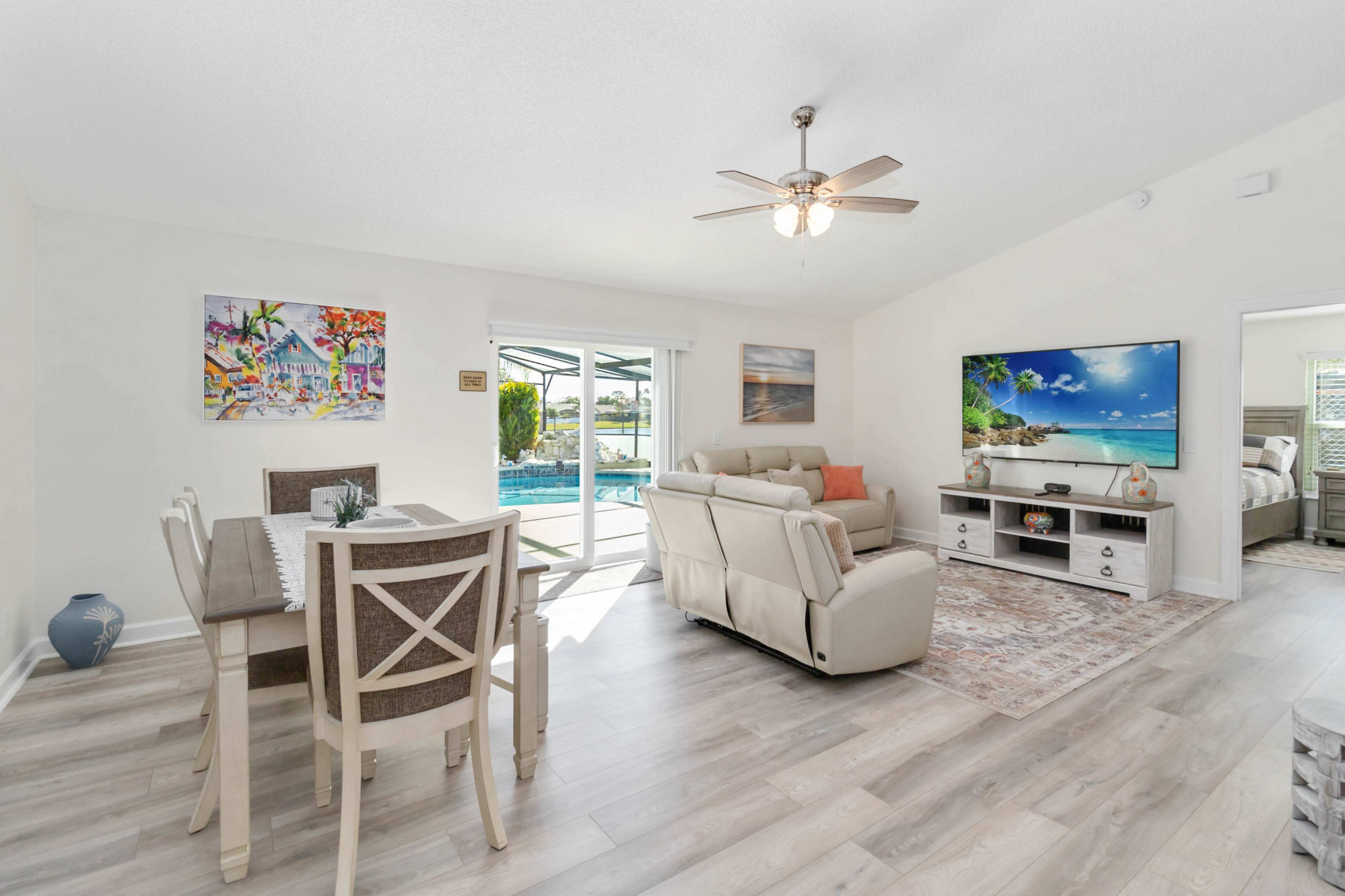 The living area features a dining table and chairs, a sectional sofa, a large television, and opens to a pool area with views of the outdoors.
