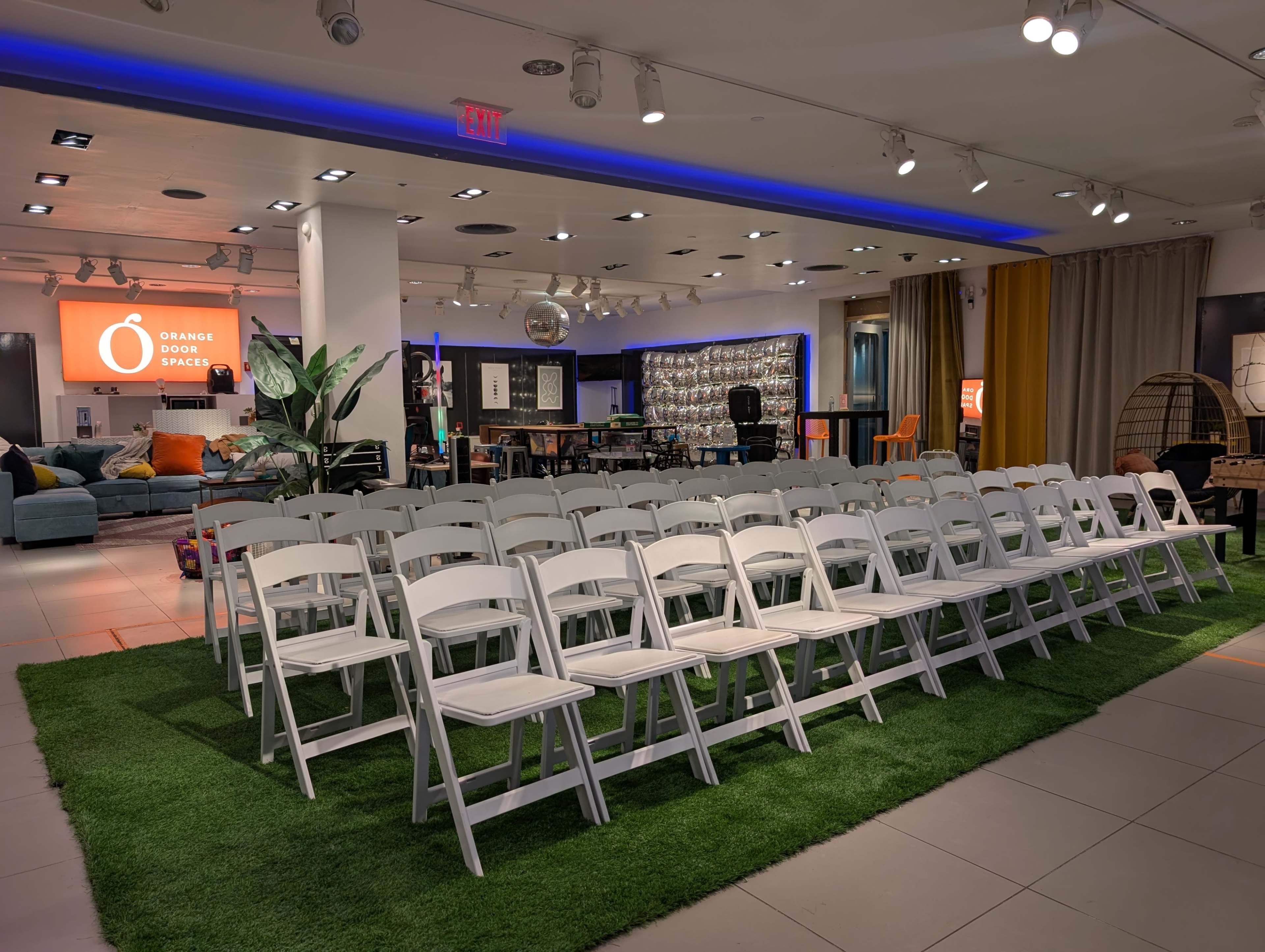 The image shows a row of empty white chairs arranged in a large indoor space with artificial grass, surrounded by colorful furniture and decorative elements.