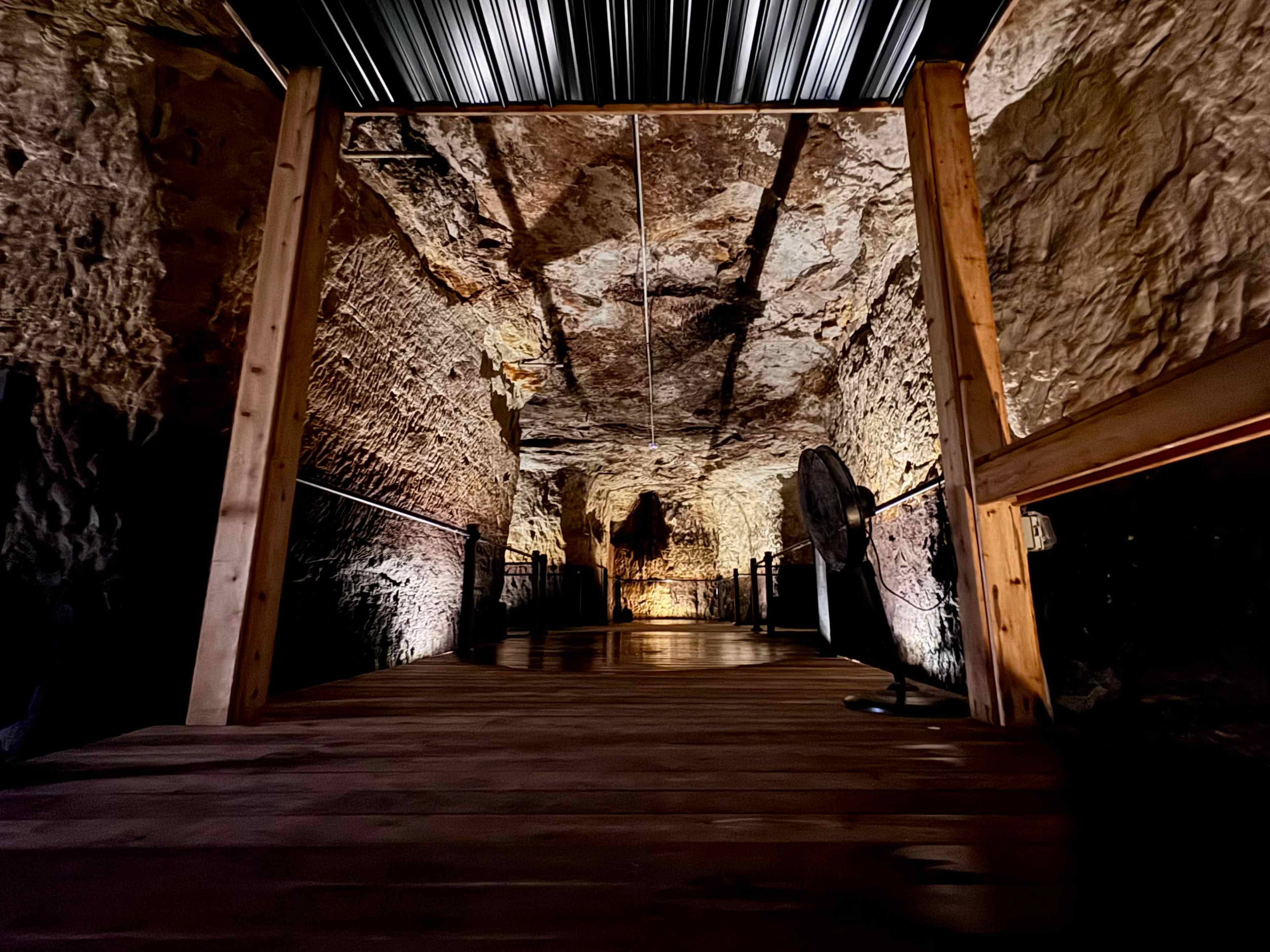 The image shows a wooden walkway leading into a cavern with illuminated rock walls and a darkened interior.