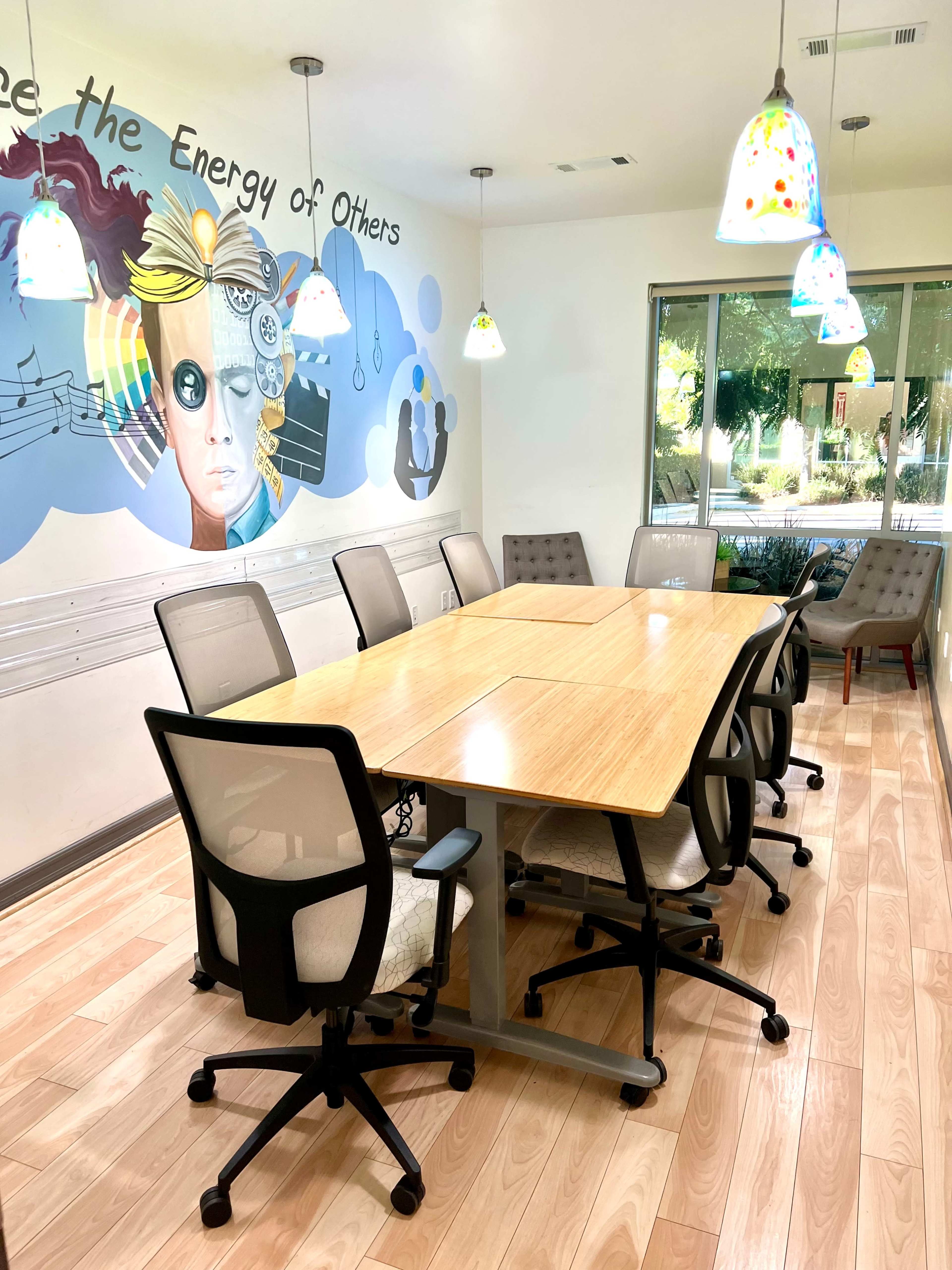 The image shows a modern conference room with a large wooden table surrounded by eight ergonomic chairs, featuring colorful pendant lights and a wall mural.