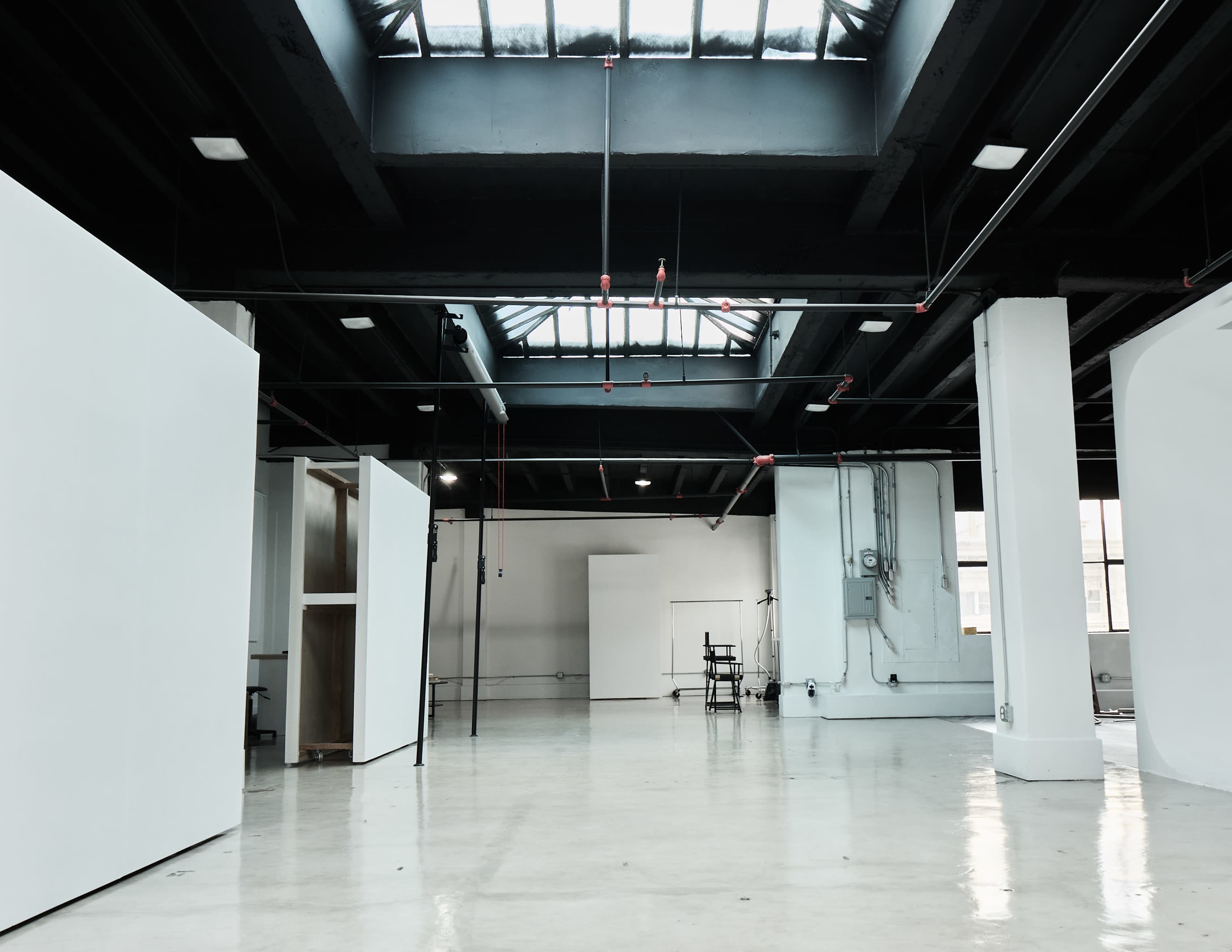 The image shows a spacious, industrial-style room with large windows, white walls, and a polished concrete floor, featuring minimal furnishings and scattered lighting.