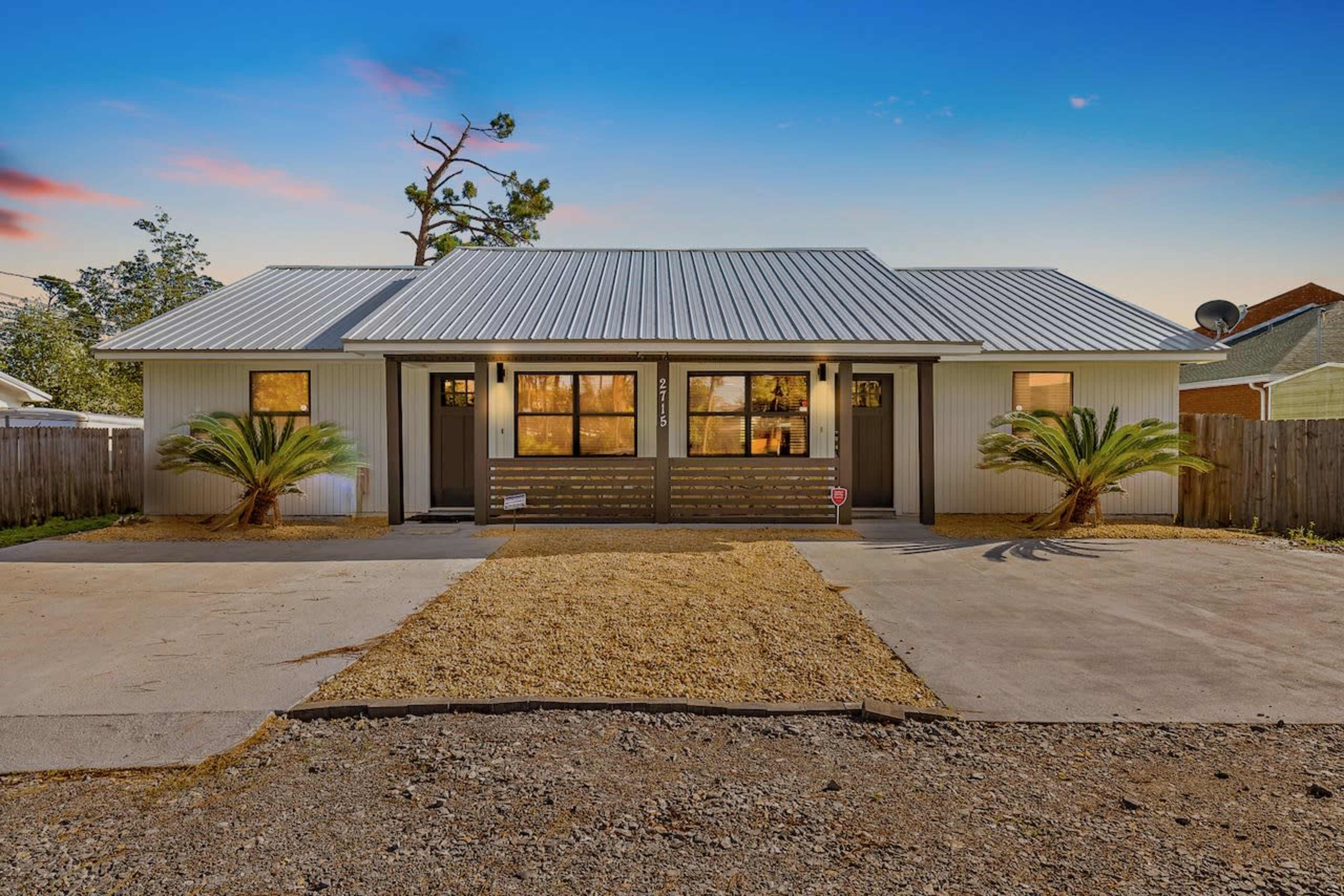 A modern, single-story duplex with a metal roof, palm trees in the front yard, and a gravel driveway.