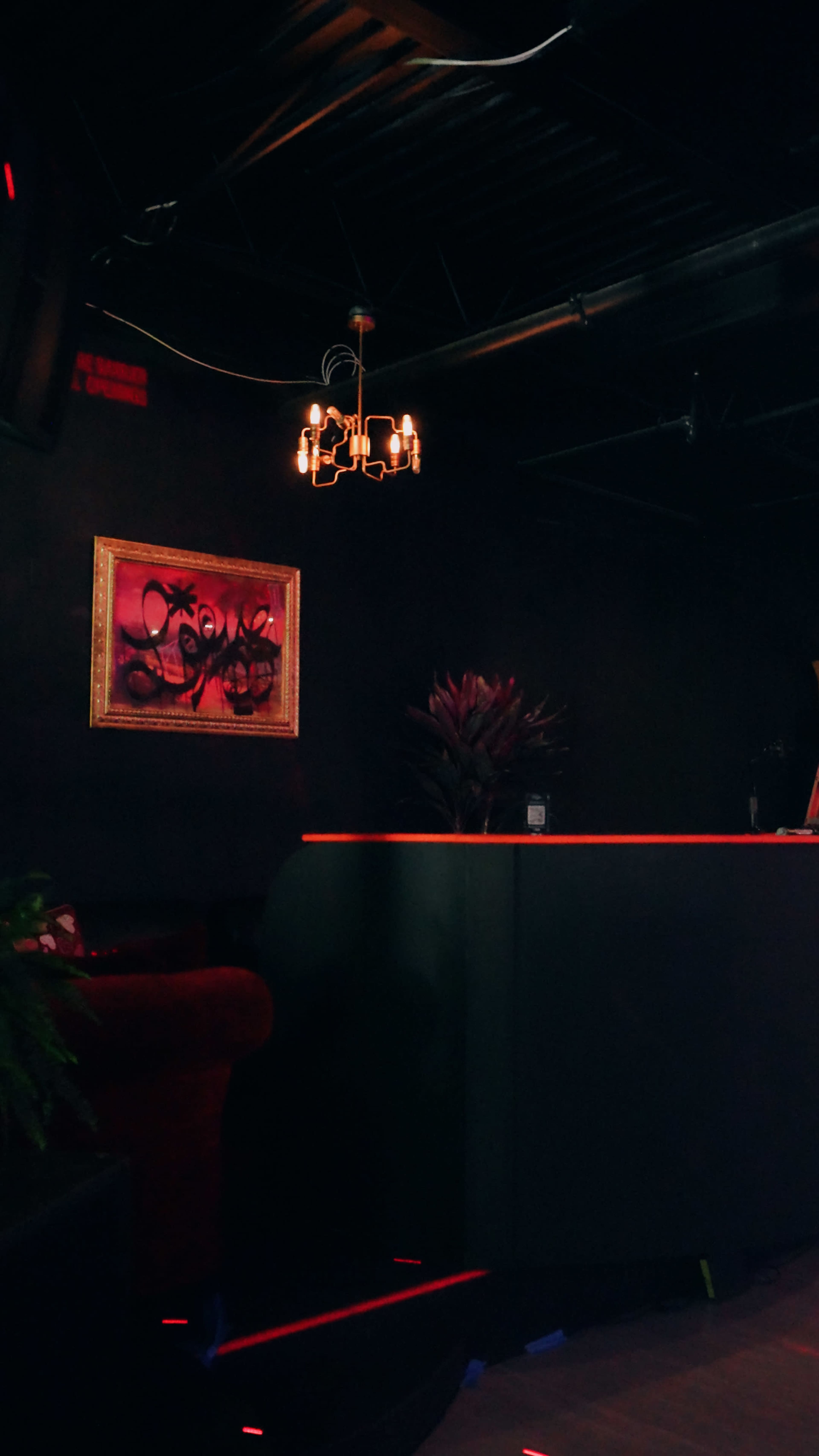 The image shows a dimly lit reception area featuring a dark wall, a framed piece of artwork, a chandelier, and a modern reception desk.