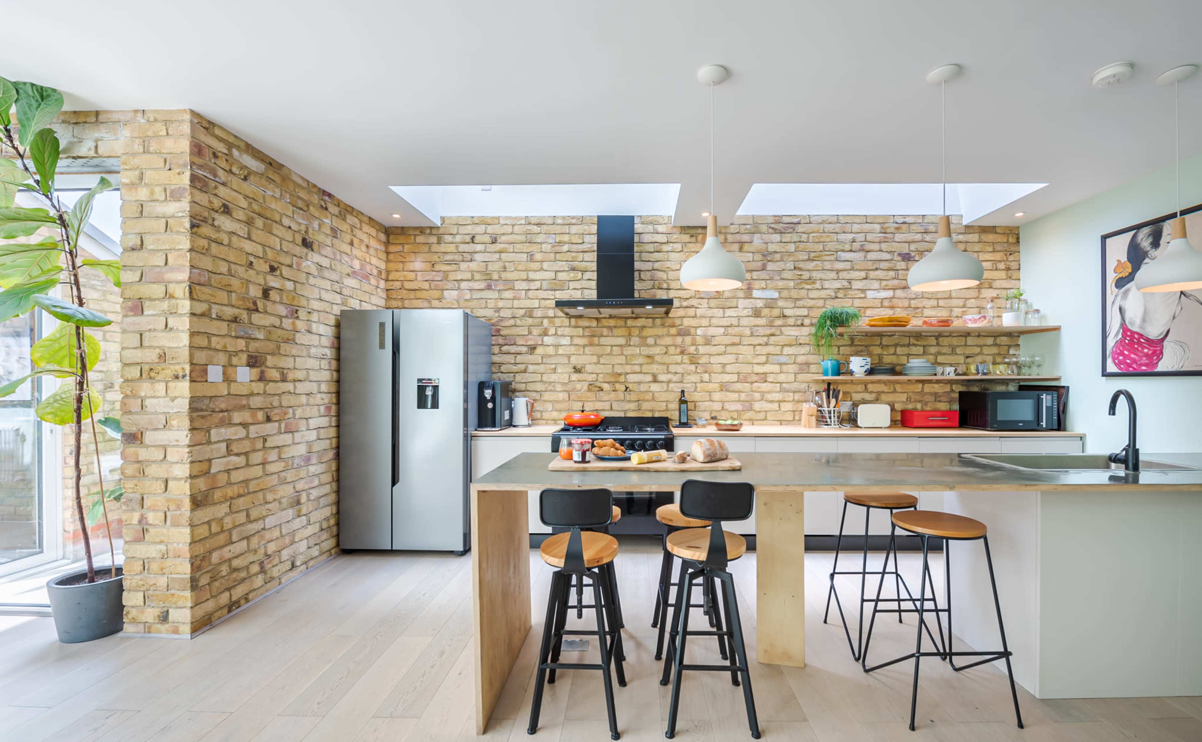 A modern kitchen features brick walls, a central island with bar stools, a stainless steel refrigerator, and large windows bringing in natural light.