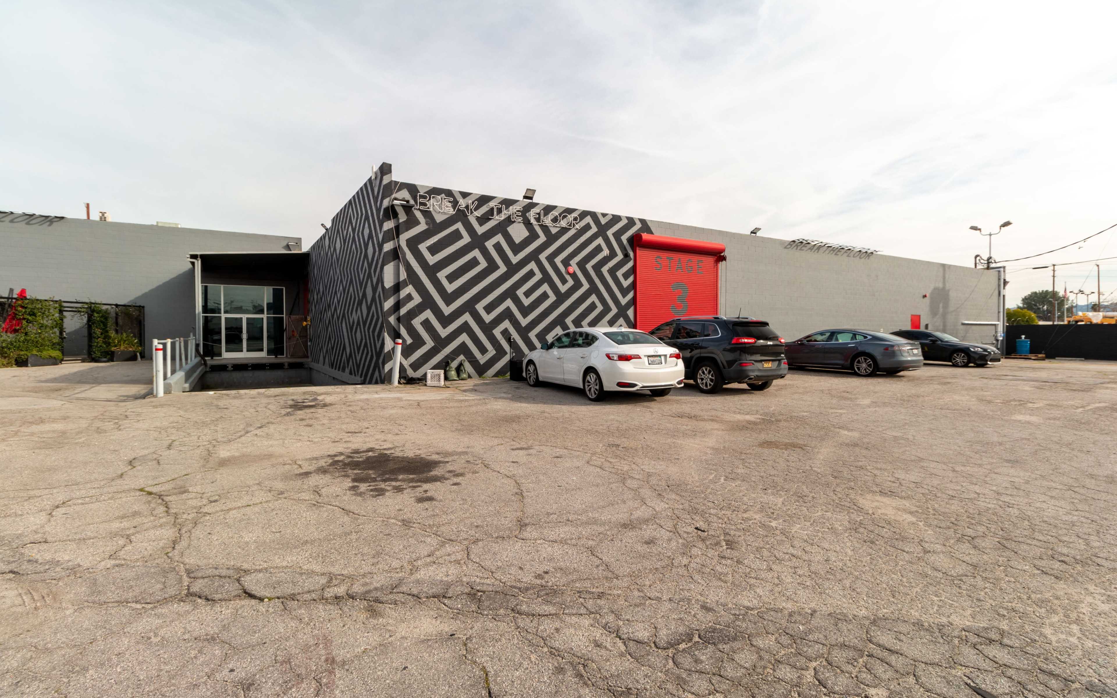 A modern building with a patterned mural on one side and several parked cars in front on a cracked asphalt surface.