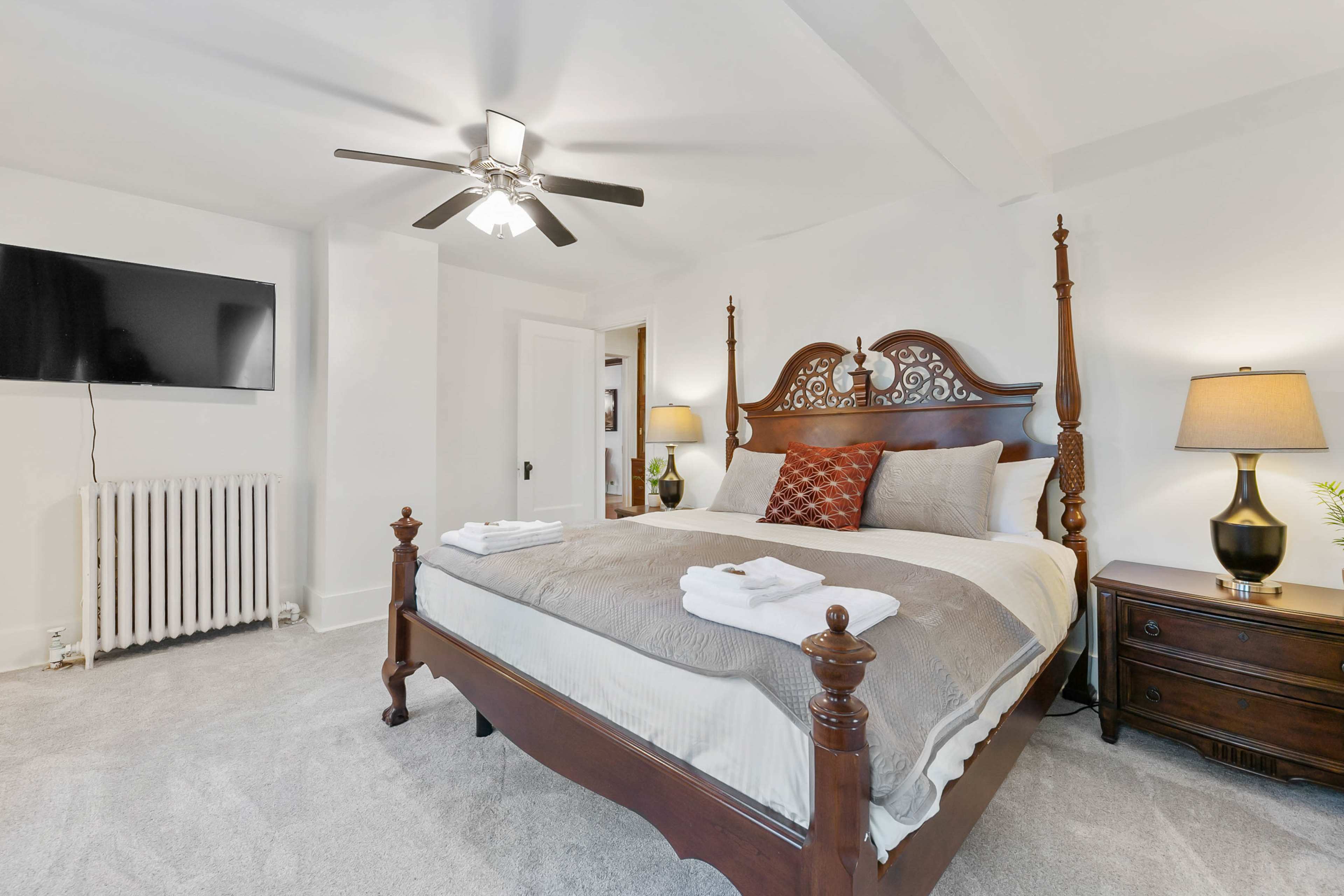 A well-furnished bedroom with a large wooden bed, bedside lamps, a television mounted on the wall, and a radiator.