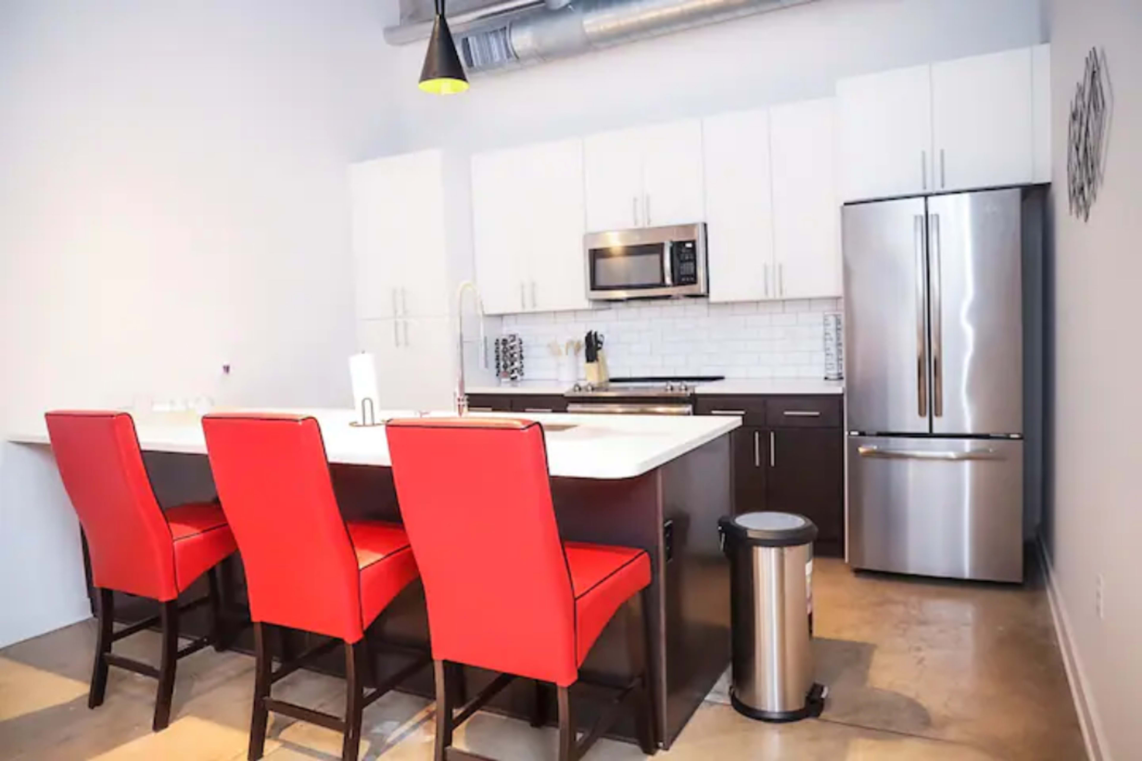 A modern kitchen with white cabinets, a stainless steel refrigerator, a microwave, and four red bar stools at a central island.