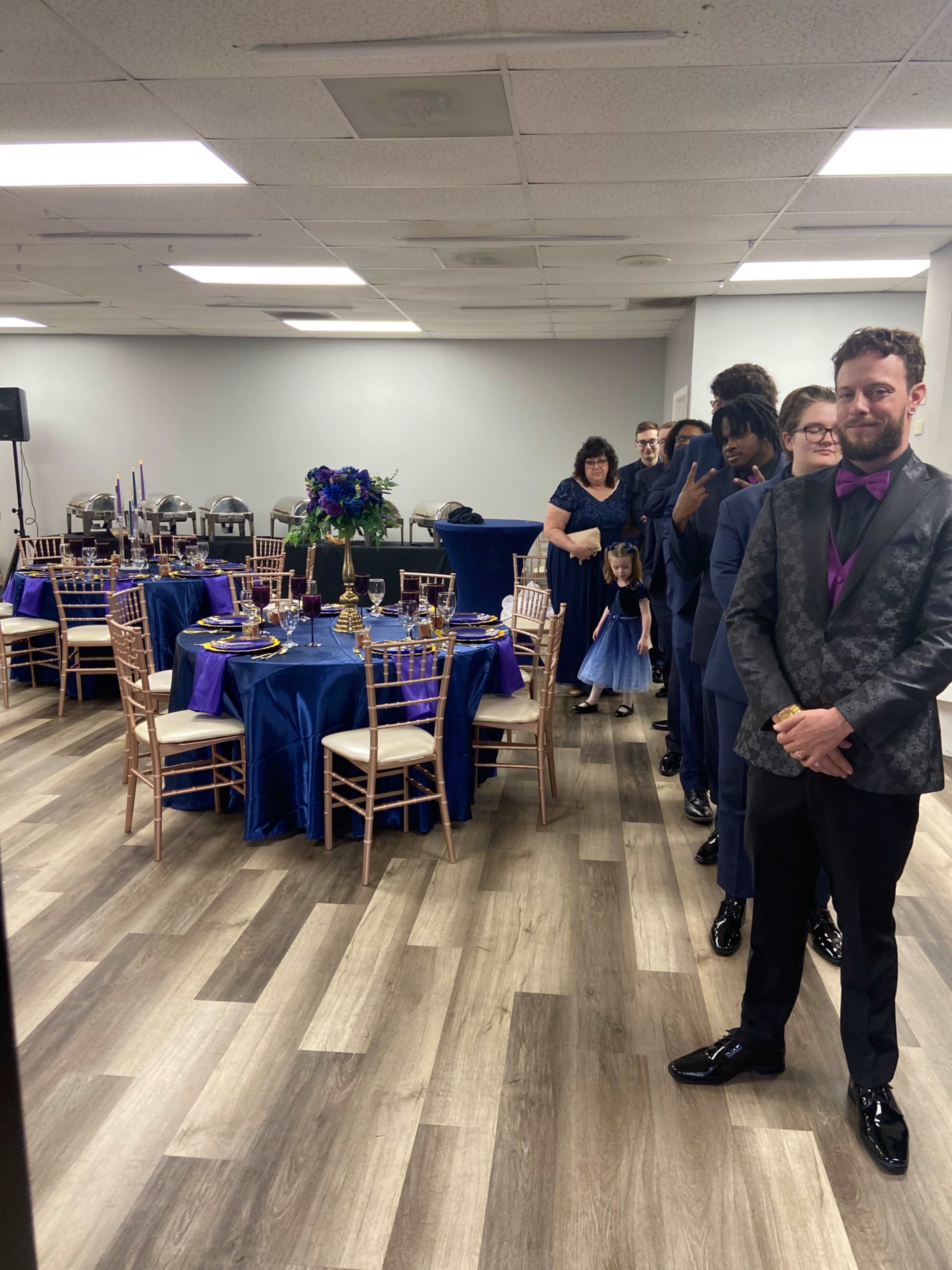 A line of well-dressed attendees stands near elegantly set tables at a banquet hall, while a small group gathers in the background.