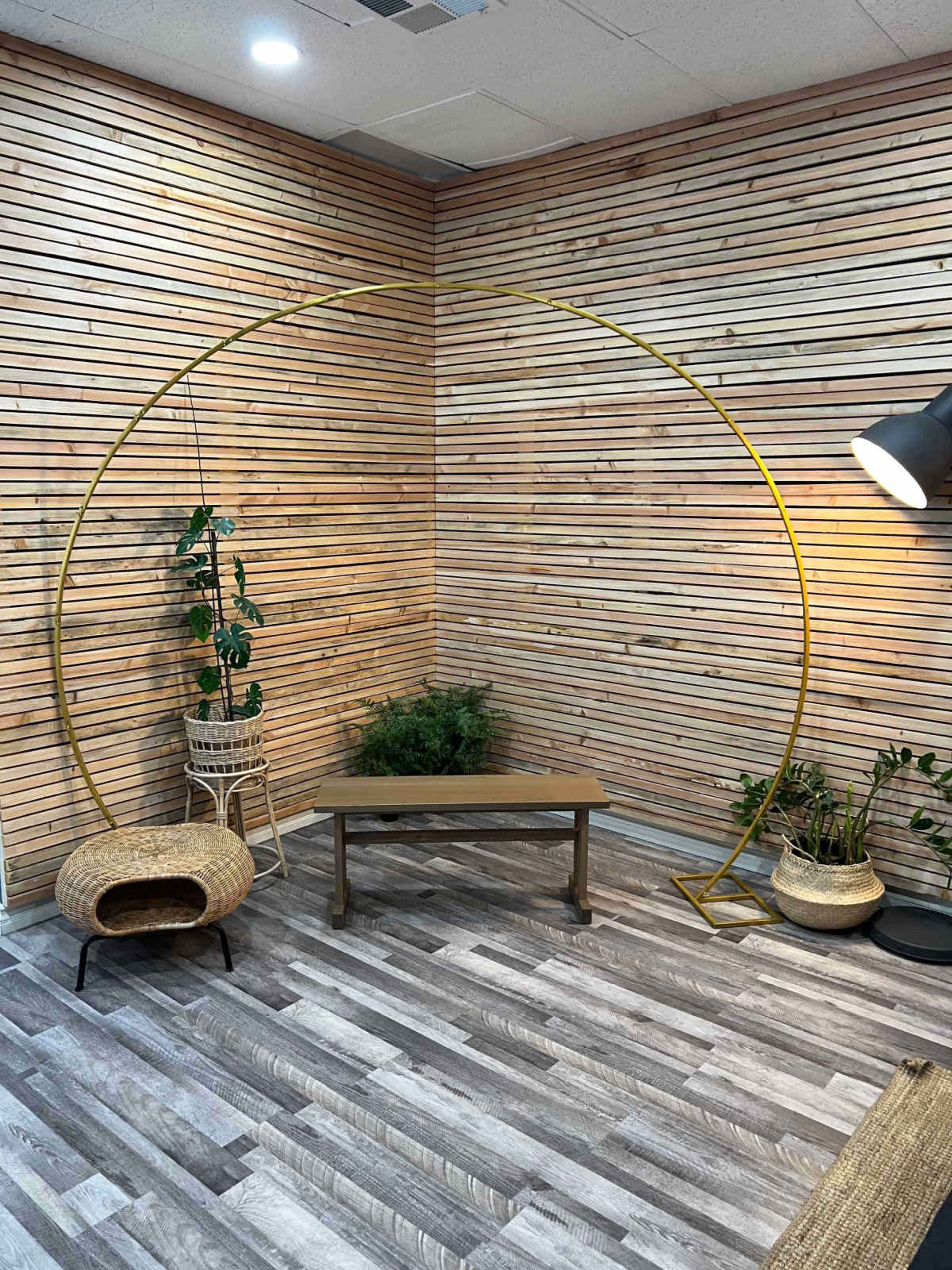 A cozy corner with a wooden slat wall, a gold circular arch, a wooden table, and various plants in woven pots.