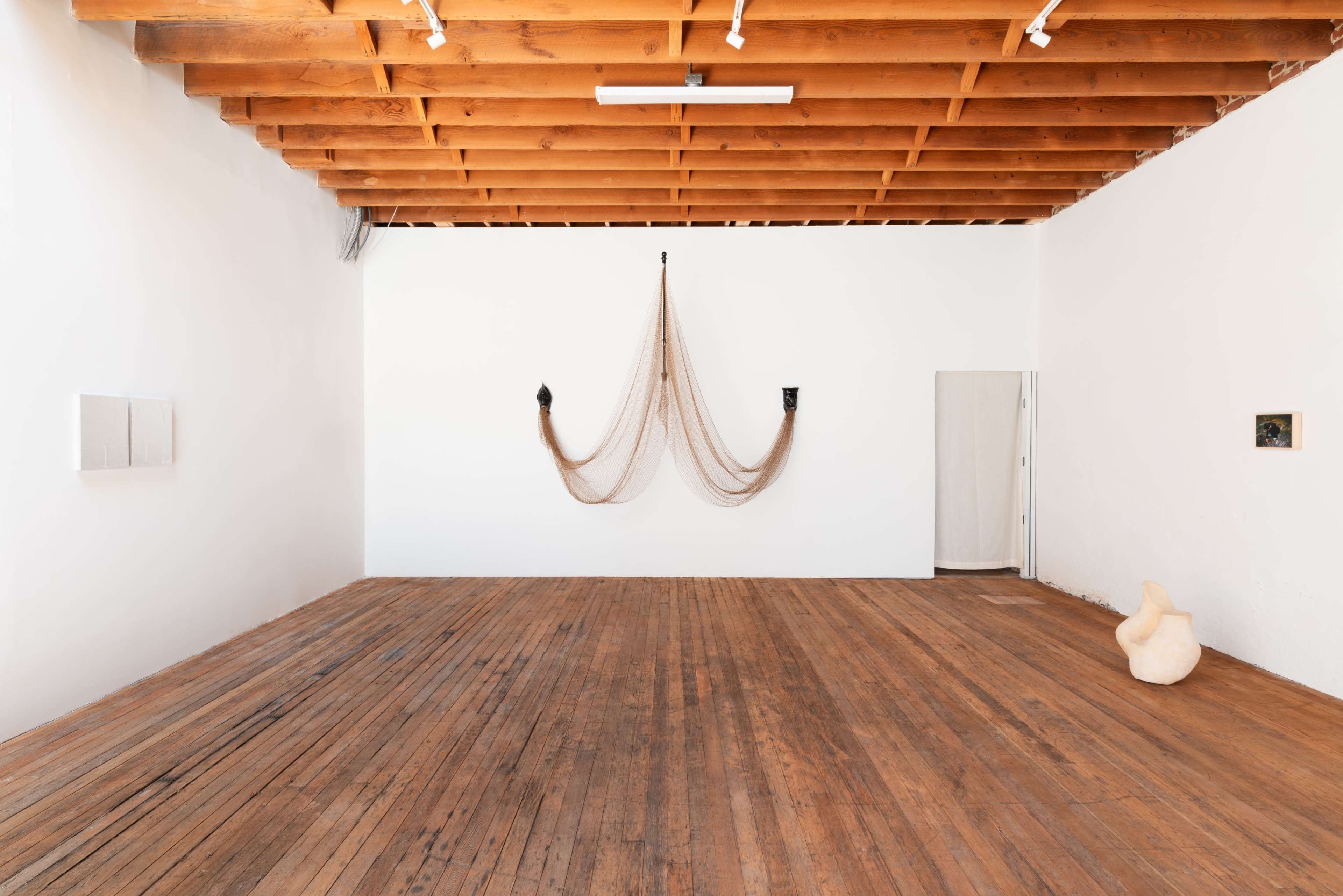 A spacious art gallery with wooden floors, white walls, and minimalist artworks, including a hanging piece made of rope and two small wall-mounted artworks.