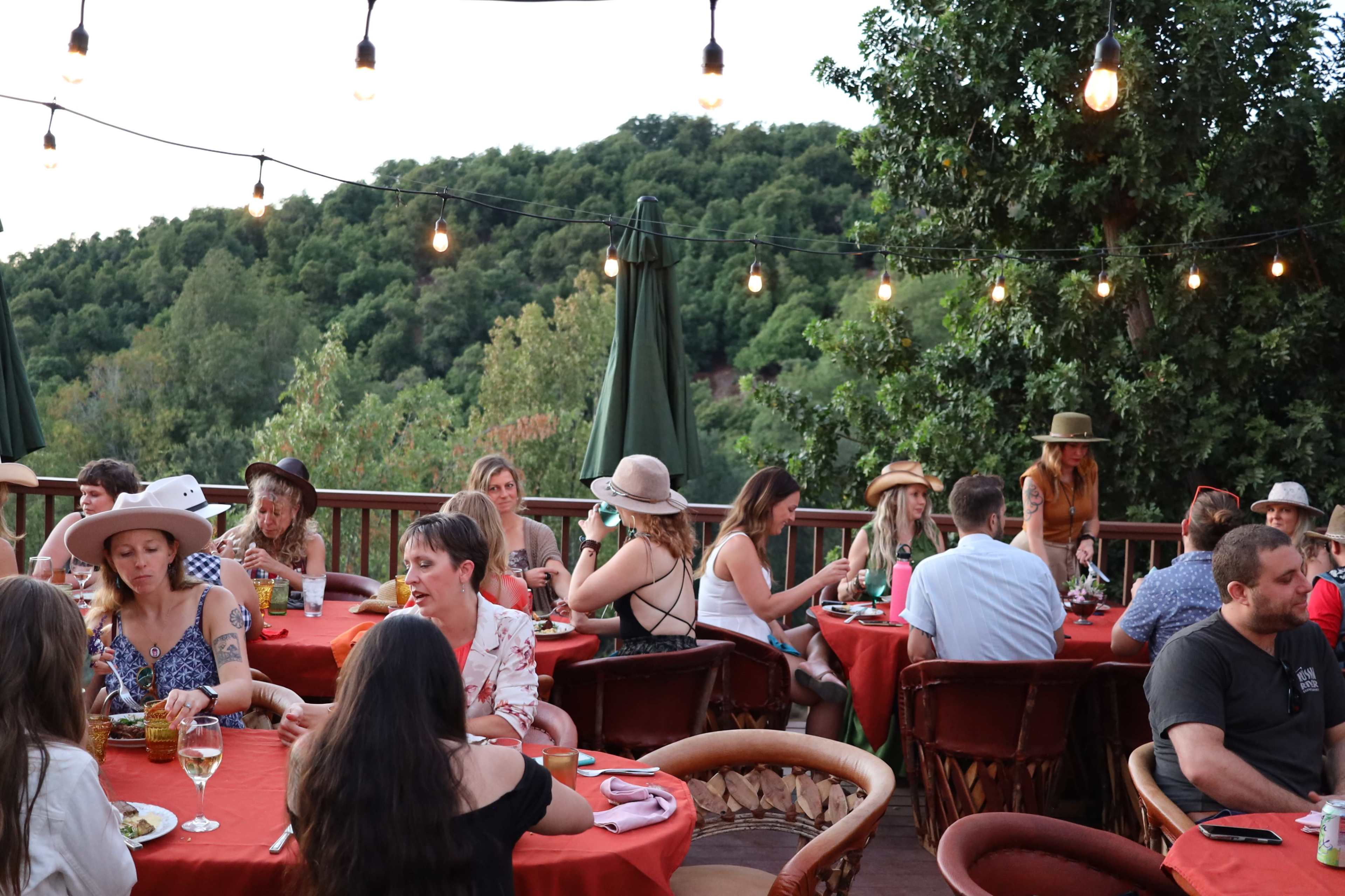 A group of people sits at tables with red tablecloths on a deck surrounded by trees, illuminated by string lights.