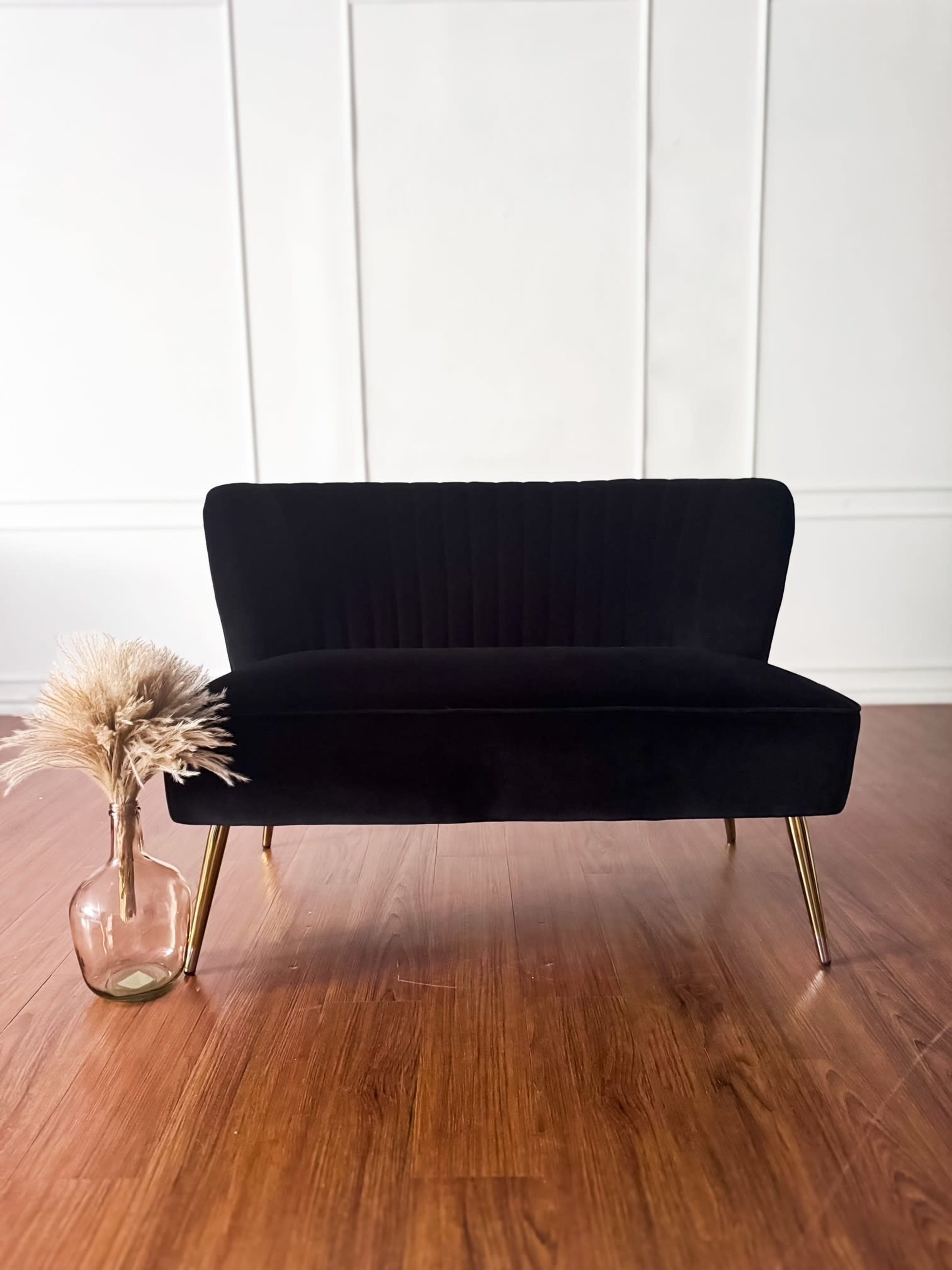 A black velvet loveseat with gold legs is positioned in front of a white wall, alongside a glass vase containing dried pampas grass.