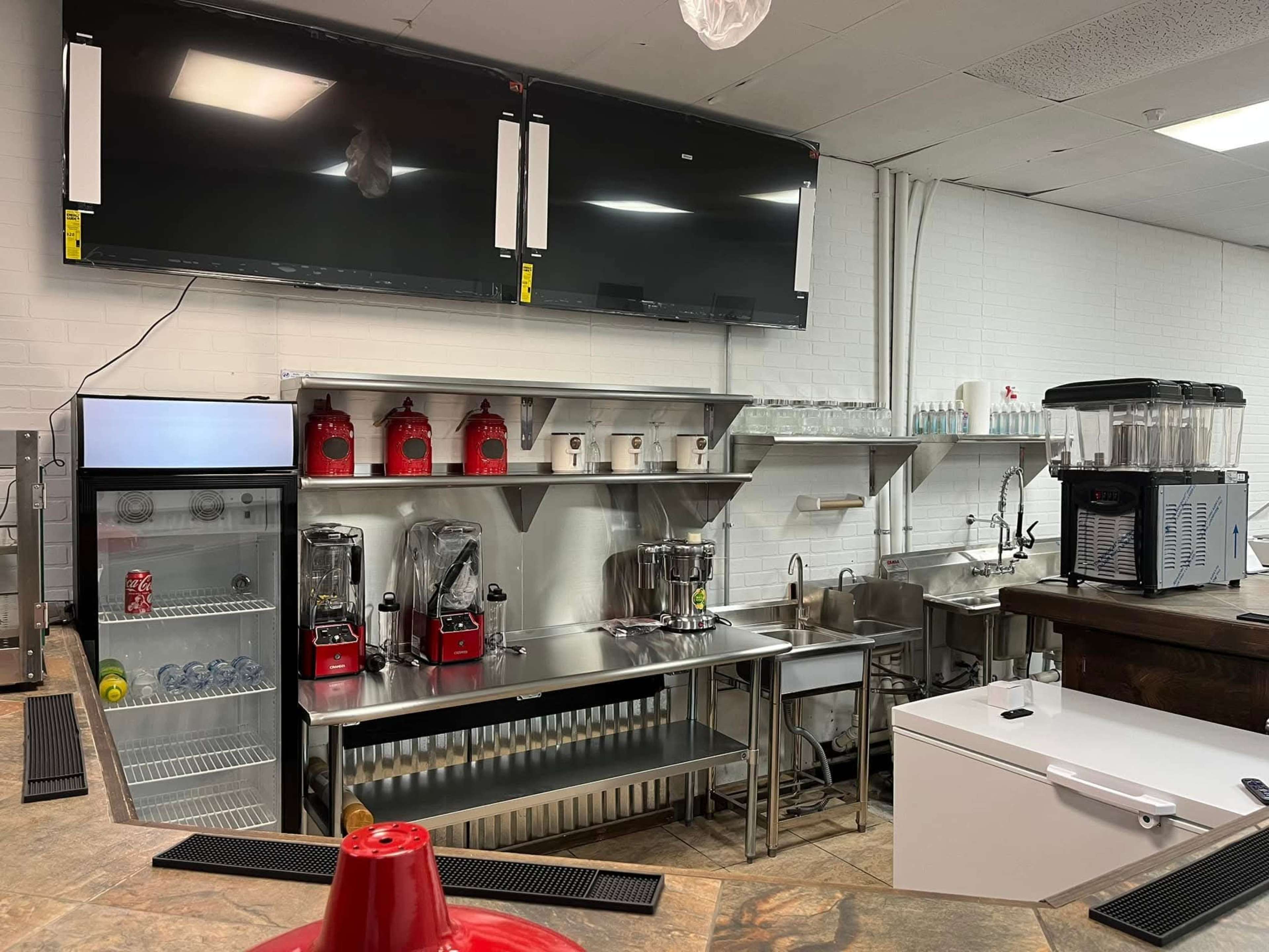 A modern commercial kitchen area features stainless steel appliances, including blenders, a refrigerator, and a drink station, with two large flat-screen TVs mounted on the wall.