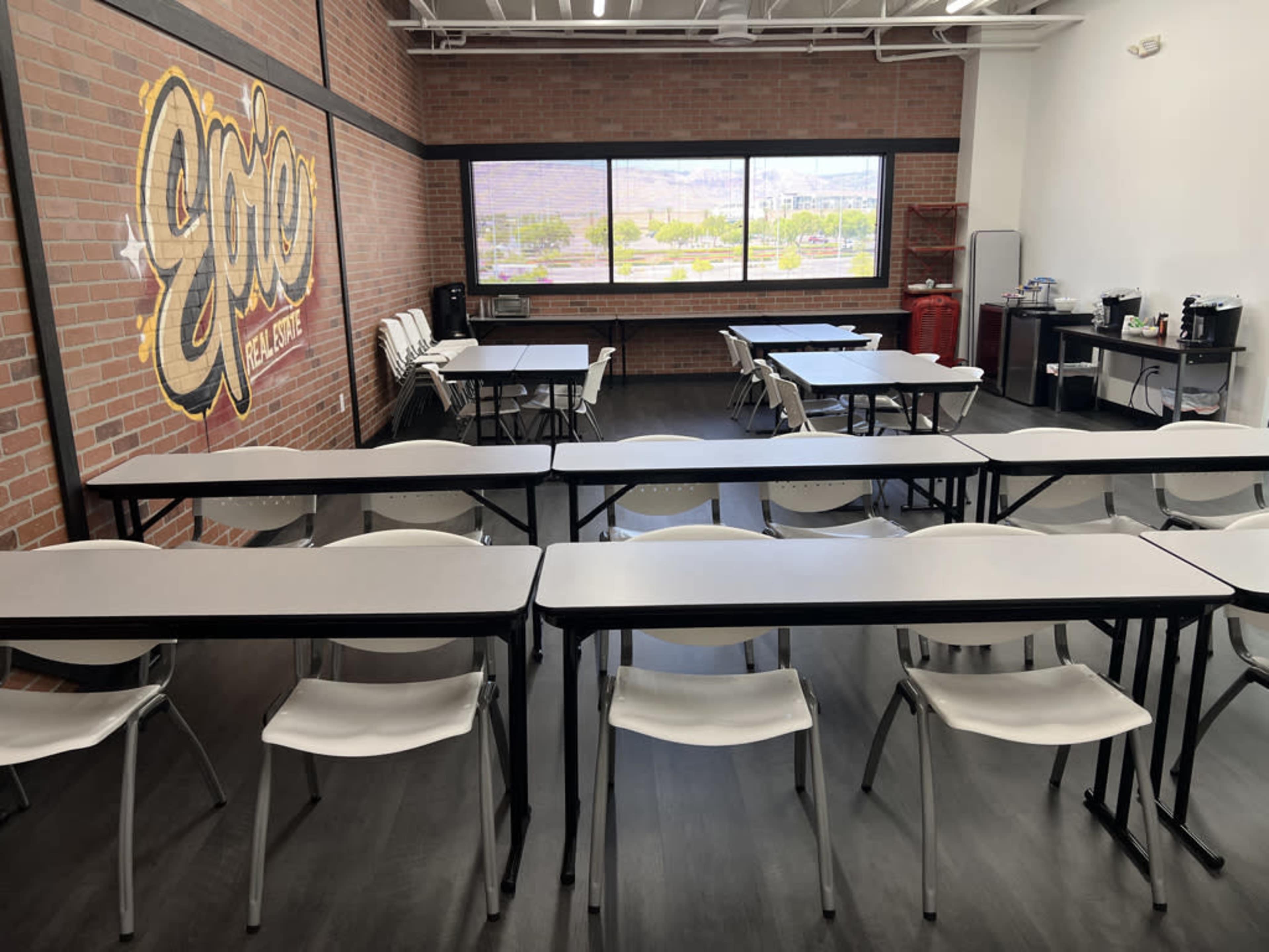 A classroom with rows of white tables and chairs facing a large window, featuring a brick wall with "Epic" graffiti.