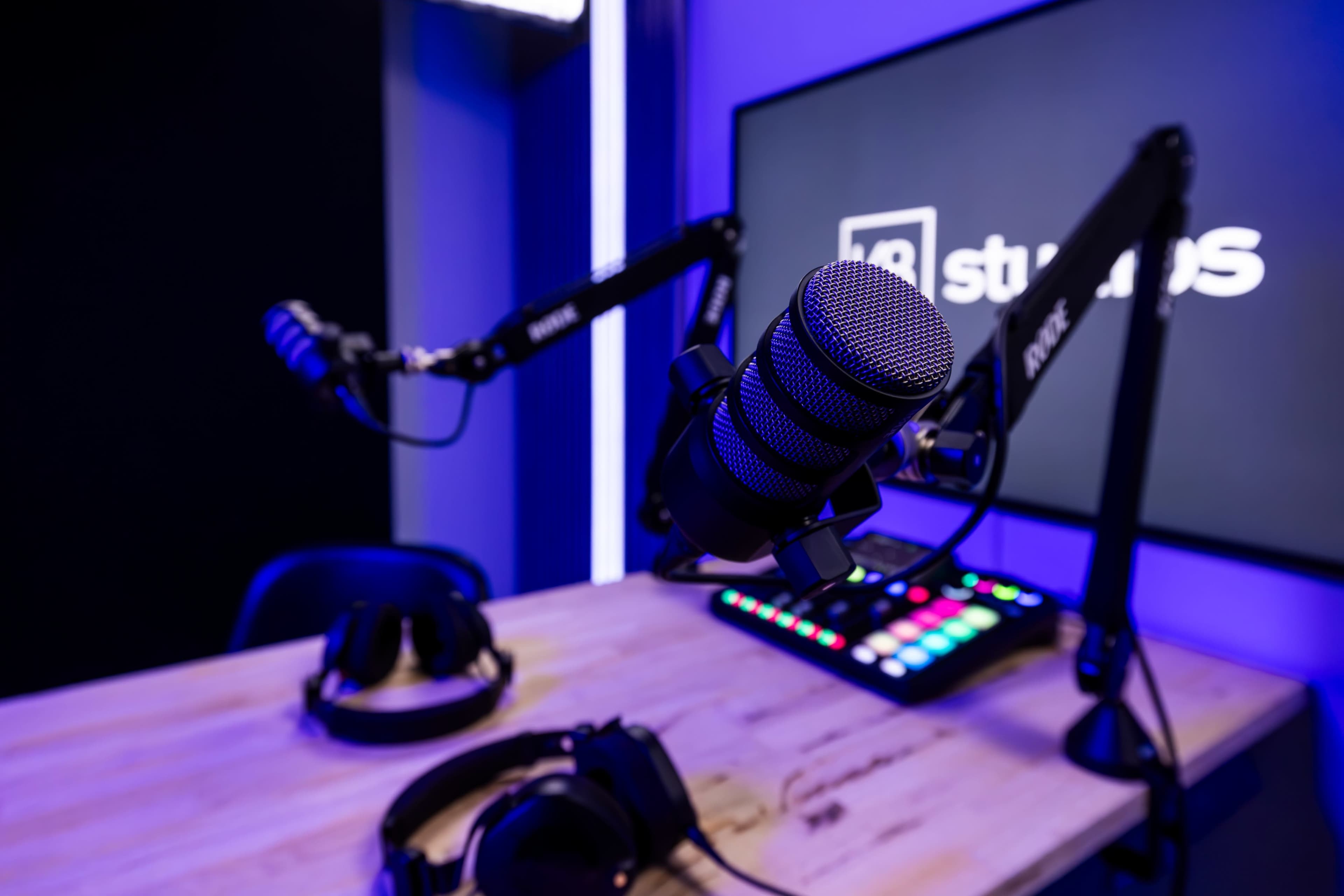 The image features a microphone mounted on an adjustable arm next to headphones, with a digital control panel visible on a wooden desk and a large screen displaying a logo in the background.