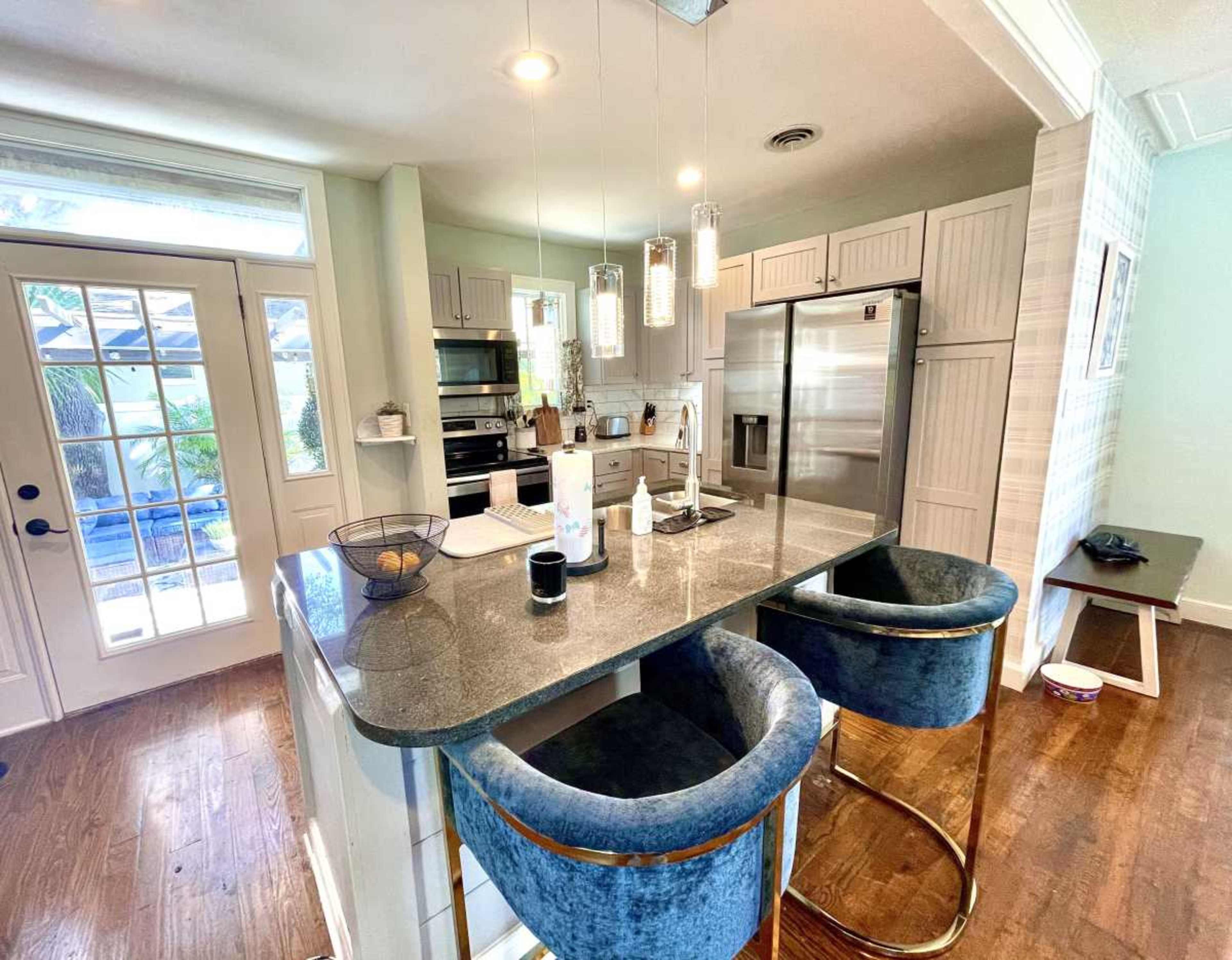 The image shows a modern kitchen featuring a dark countertop, bar stools, stainless steel appliances, and a door leading to a patio.