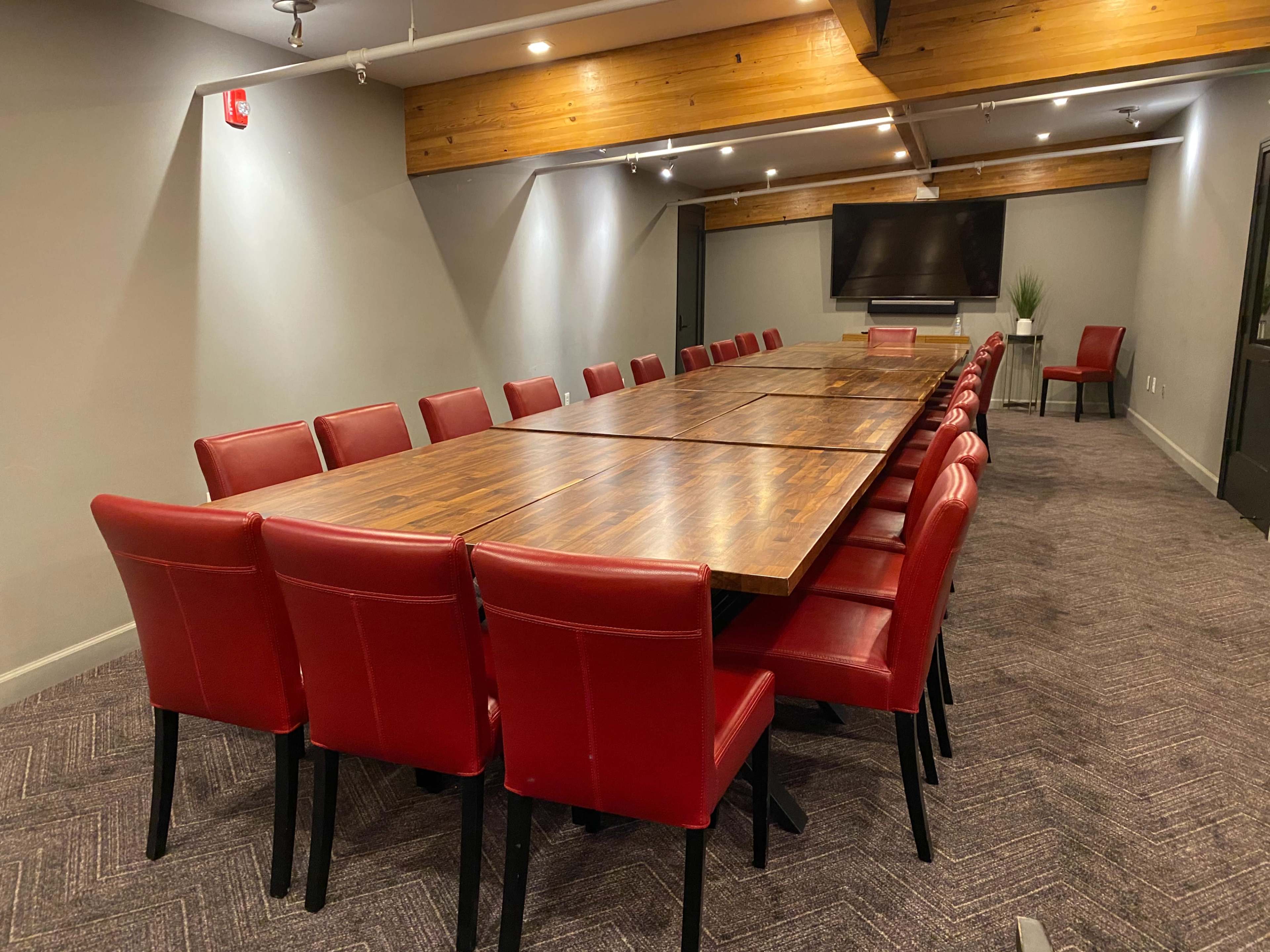 A long conference table surrounded by red chairs is situated in a meeting room with gray walls and wooden accents.