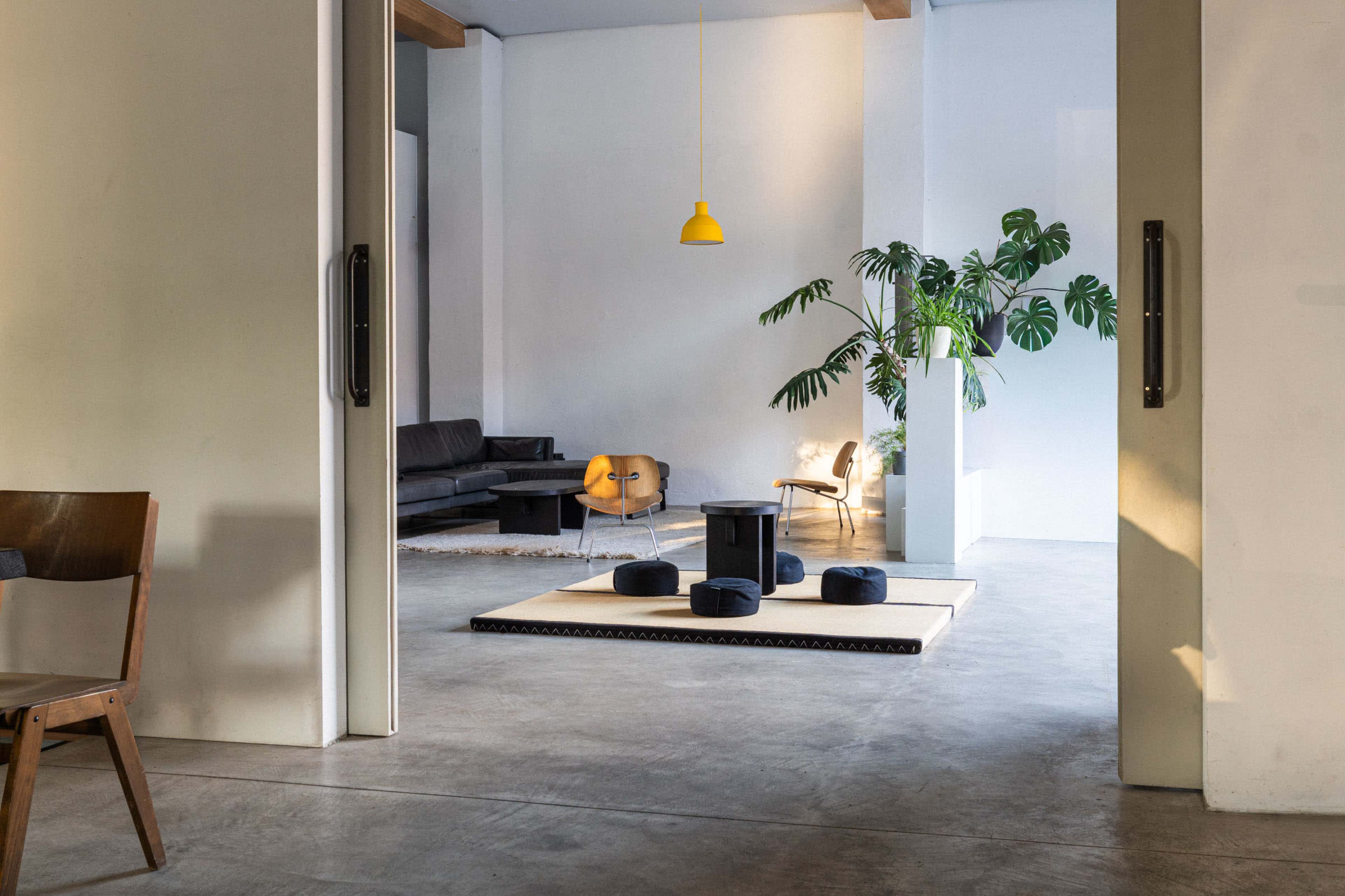 The image shows a bright, modern interior space with a seating area featuring a sofa, chairs, and a potted plant, along with a low table surrounded by cushions on a mat.