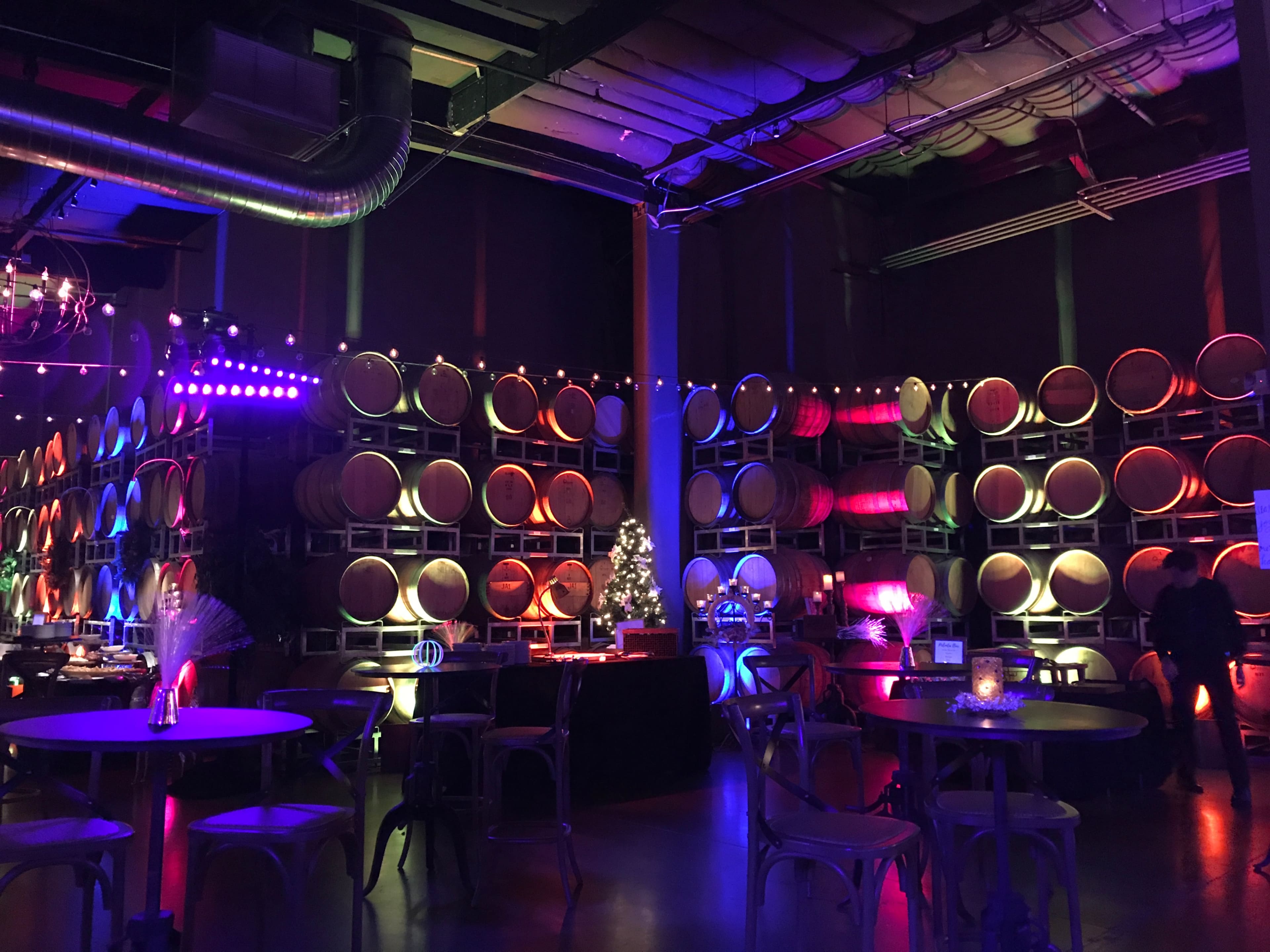 The scene features a dimly lit winery with rows of wine barrels stacked against the wall, illuminated by colorful lights and decorated for an event.