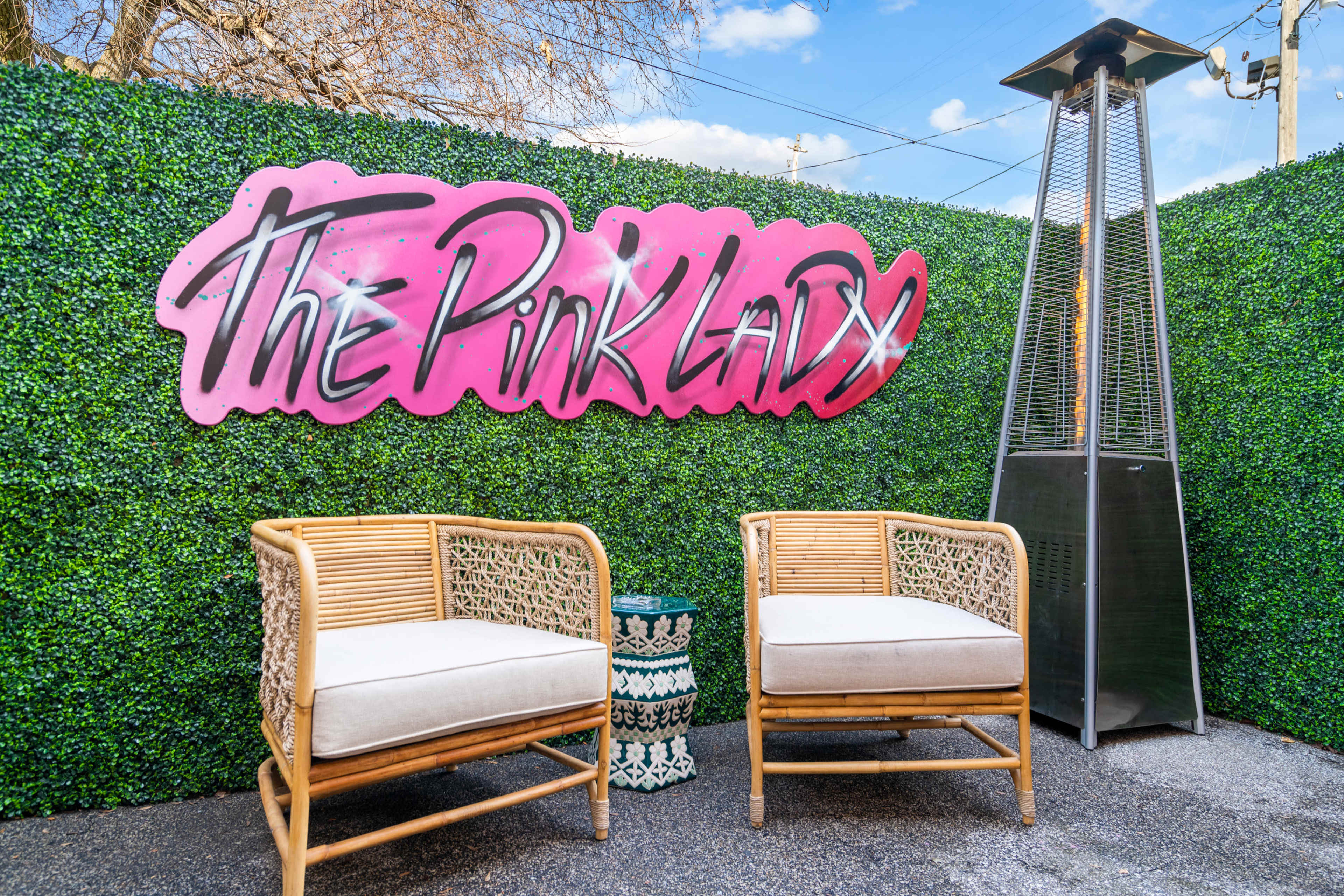 A vibrant sign reading "The Pink Lady" is displayed against a green hedge, accompanied by two wicker chairs and a patio heater.