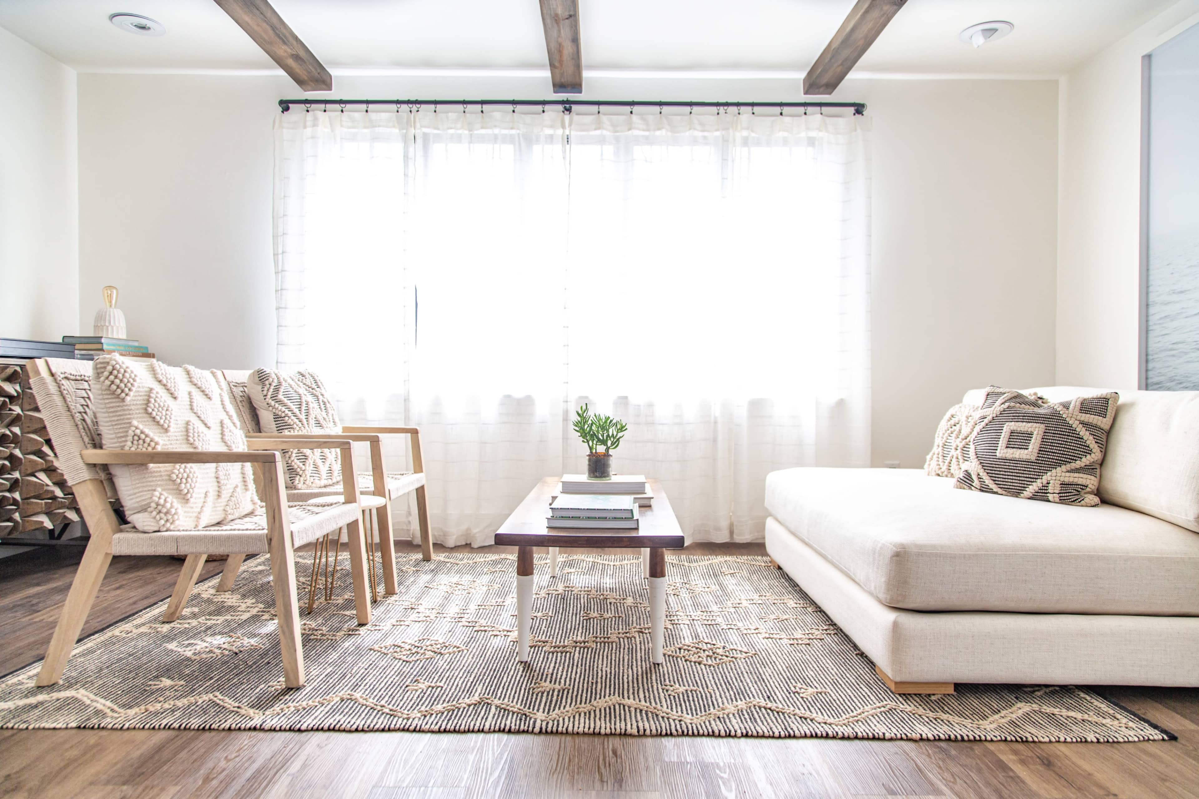 A modern living room features a light-colored sofa, wooden chairs, a coffee table, and a large window with sheer curtains.