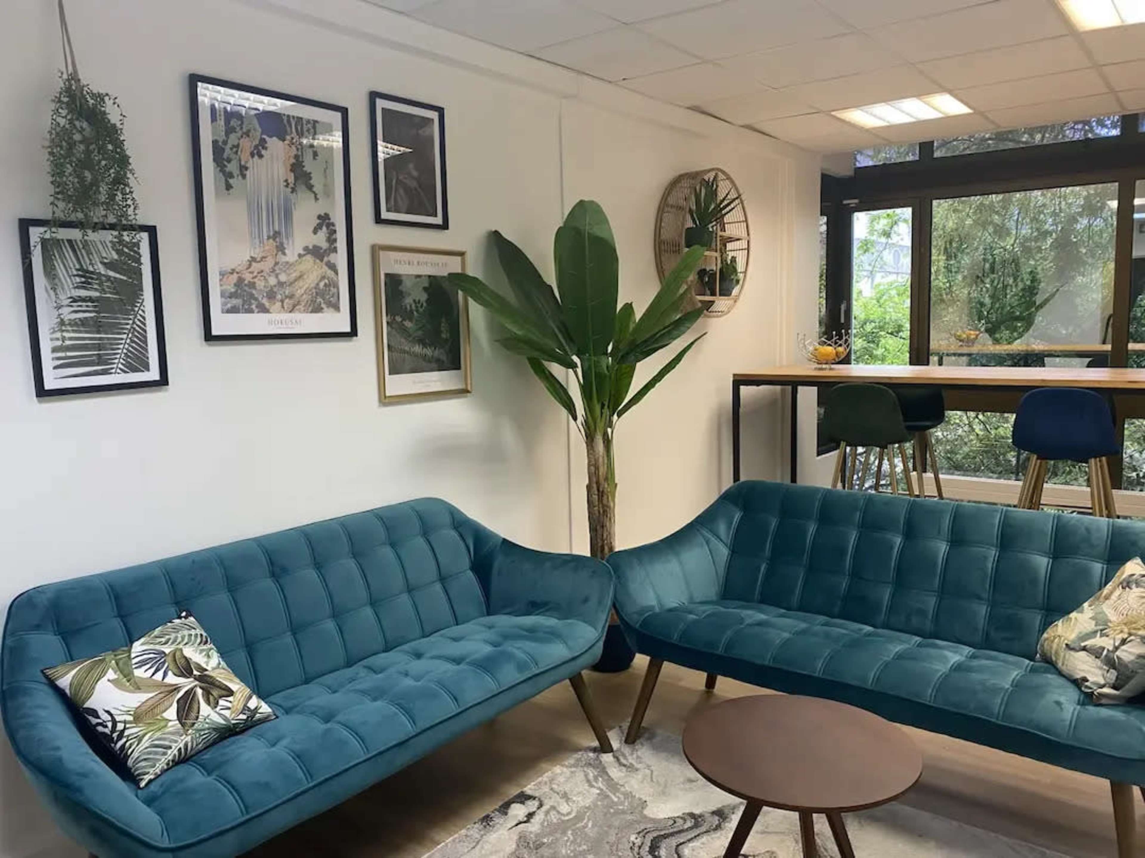 The image features a modern lounge area with two teal sofas, a circular coffee table, and framed artwork on the walls, surrounded by plants and large windows.