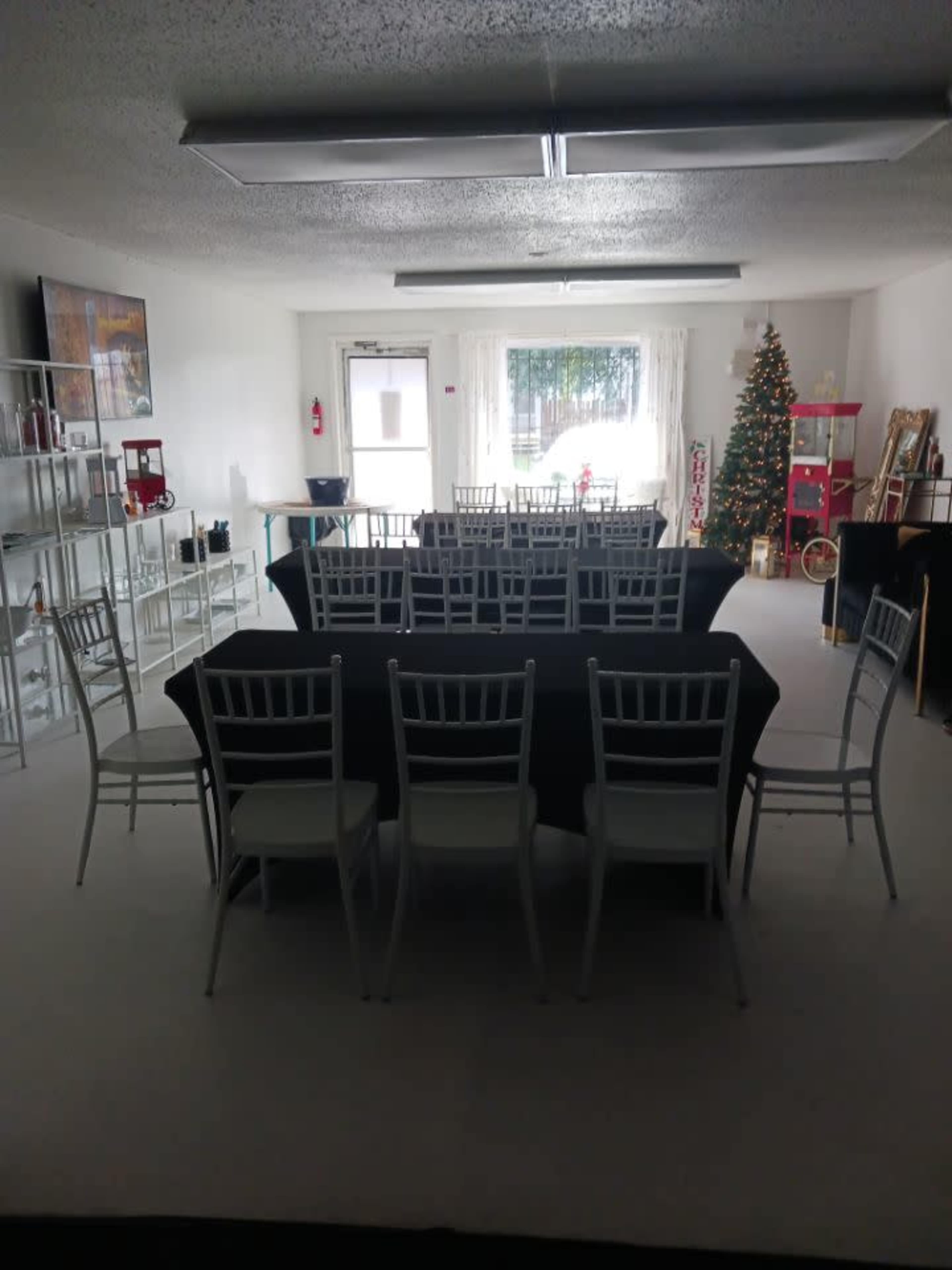 A spacious room set up for an event, featuring multiple tables covered with black cloth and surrounded by white chairs, with a decorated Christmas tree in one corner.