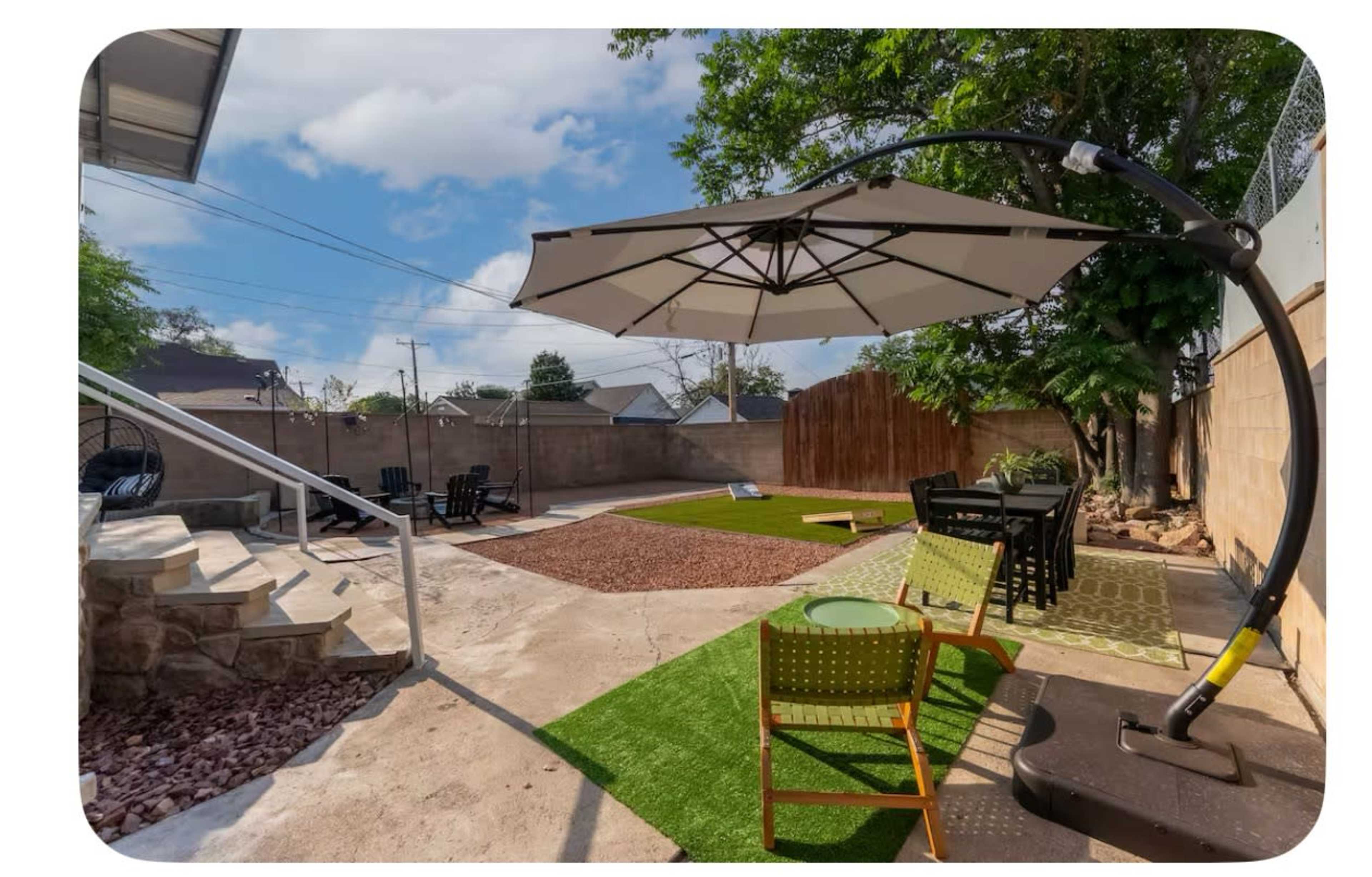 The image shows a backyard with a patio area featuring a large umbrella, artificial grass, gravel, and seating arrangements, surrounded by a wall and trees.