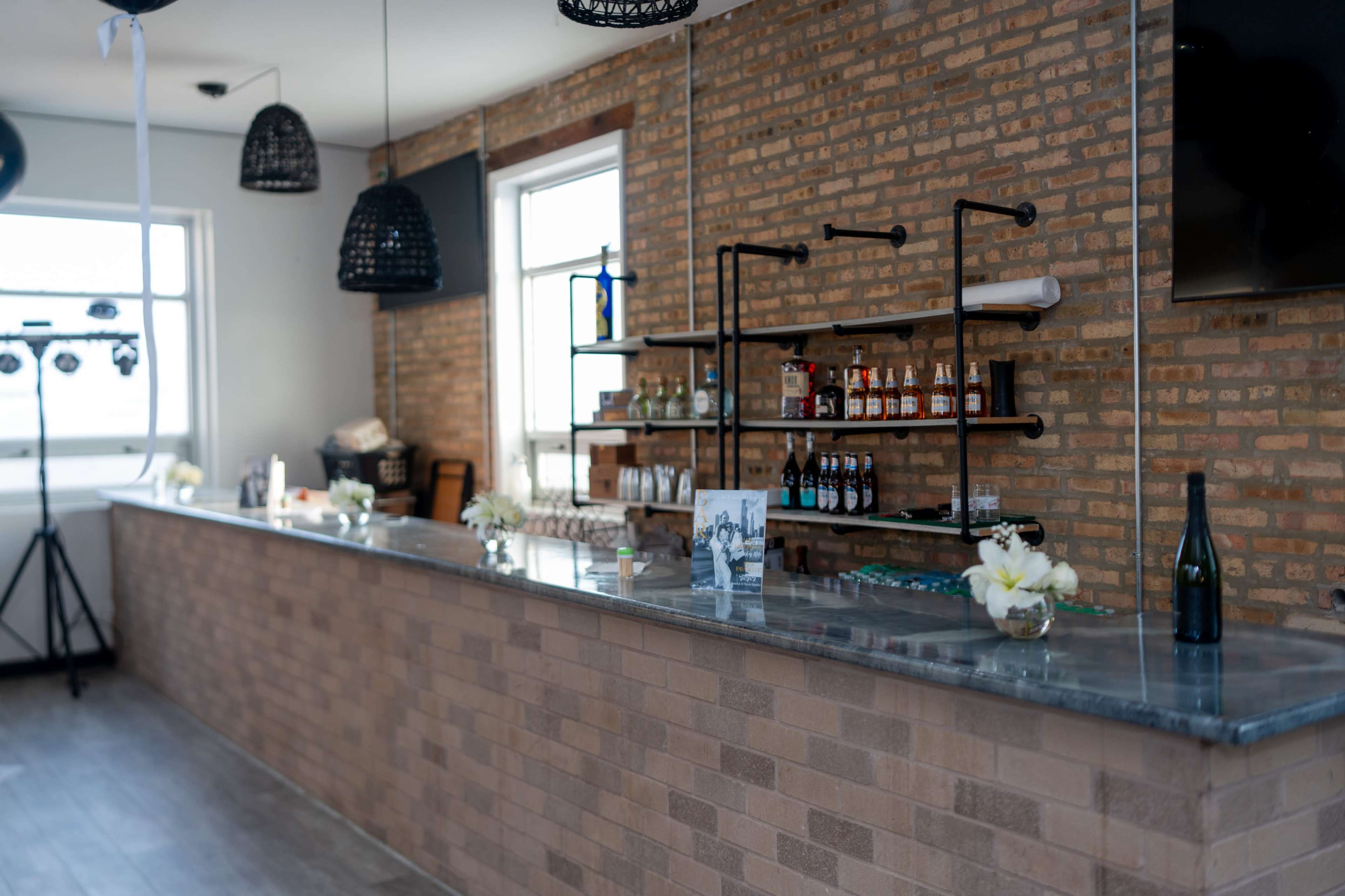 The image shows a modern bar with a brick wall, a sleek countertop, and various bottles displayed on shelves.