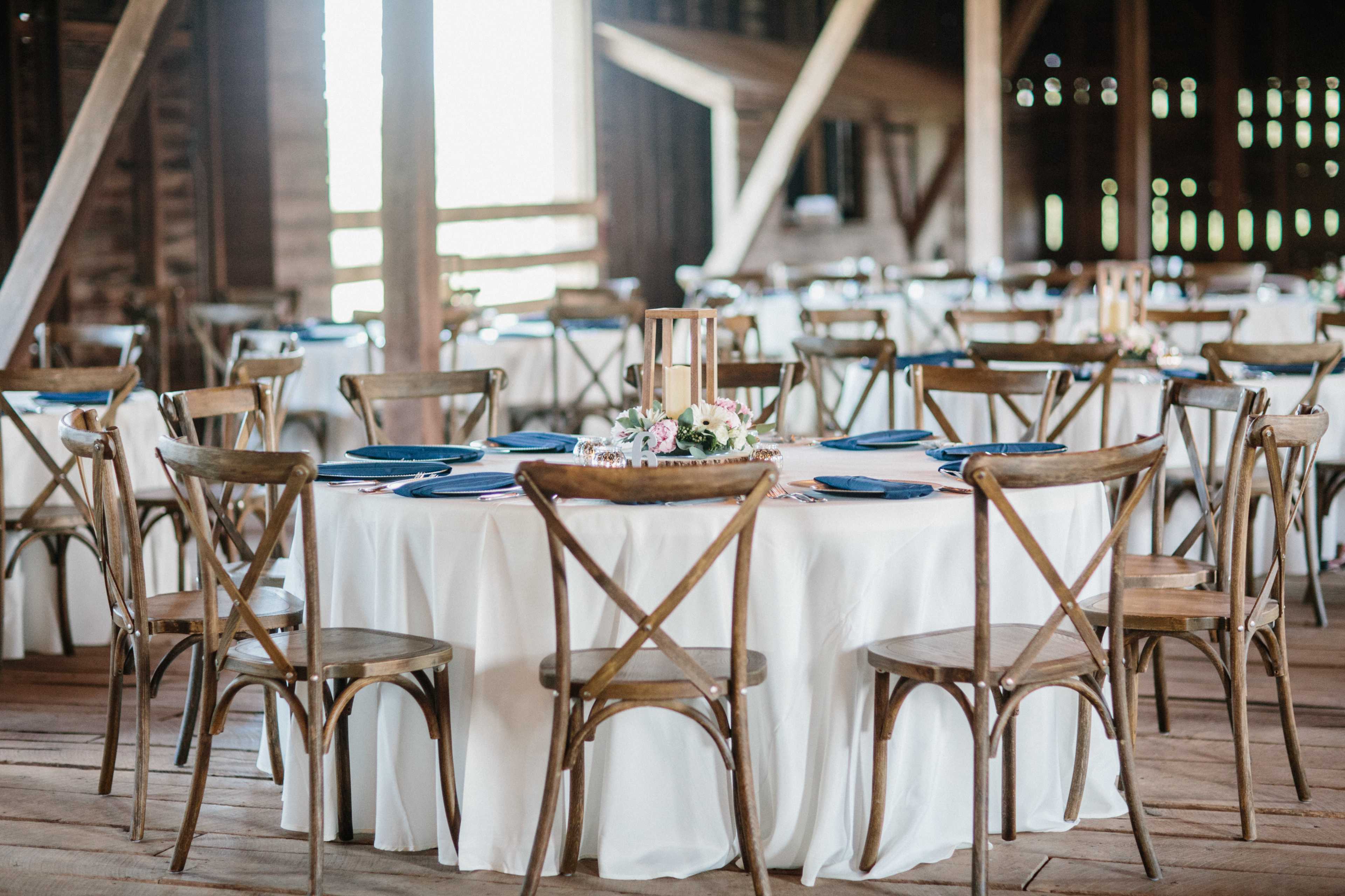 A spacious barn interior features round tables with white tablecloths, wooden chairs, and decorative centerpieces.