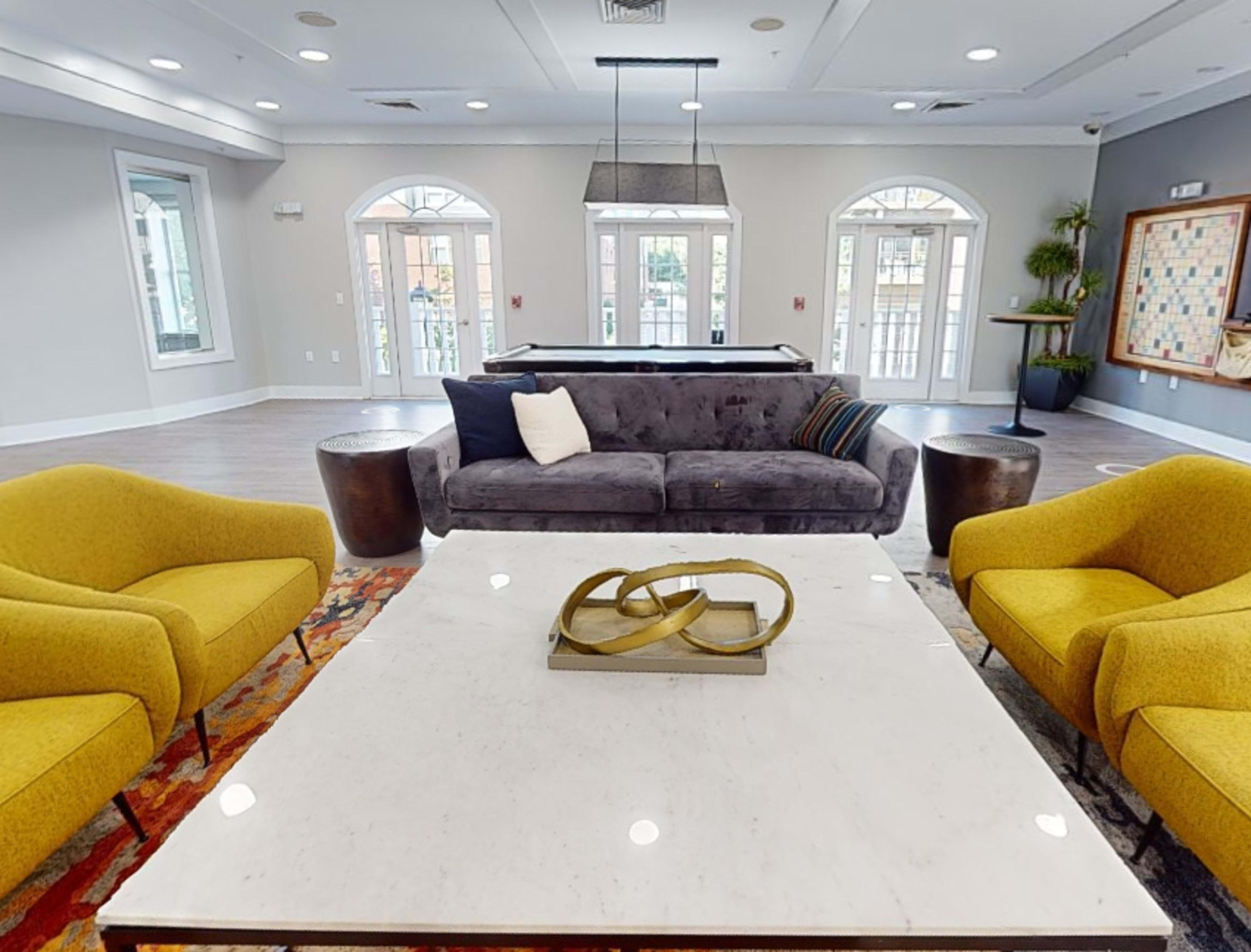 A modern lounge area featuring a gray sofa, two yellow armchairs, a marble coffee table, and large windows letting in natural light.