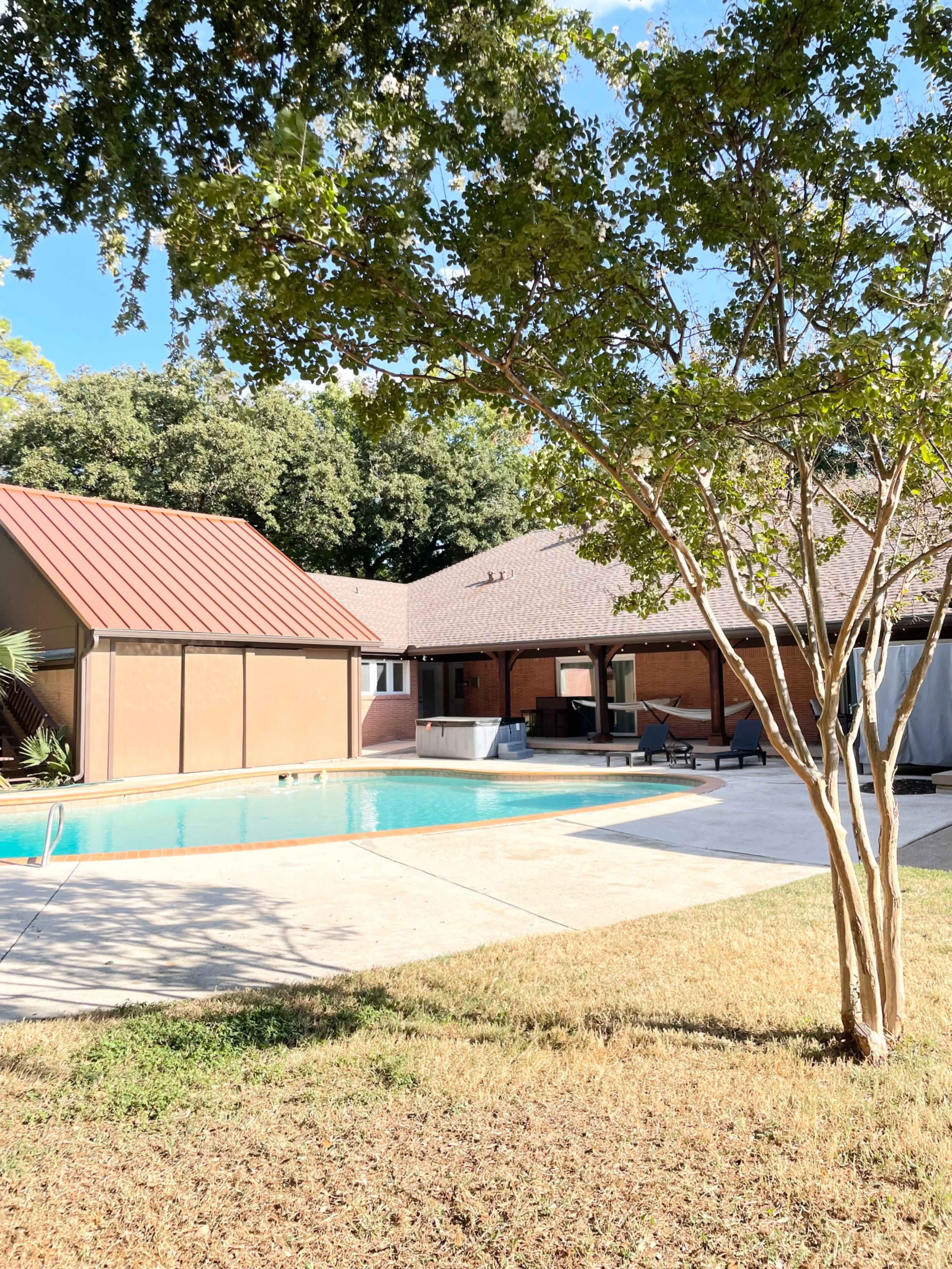A residential backyard features a swimming pool adjacent to a single-story house with a red metal roof and a tree in the foreground.