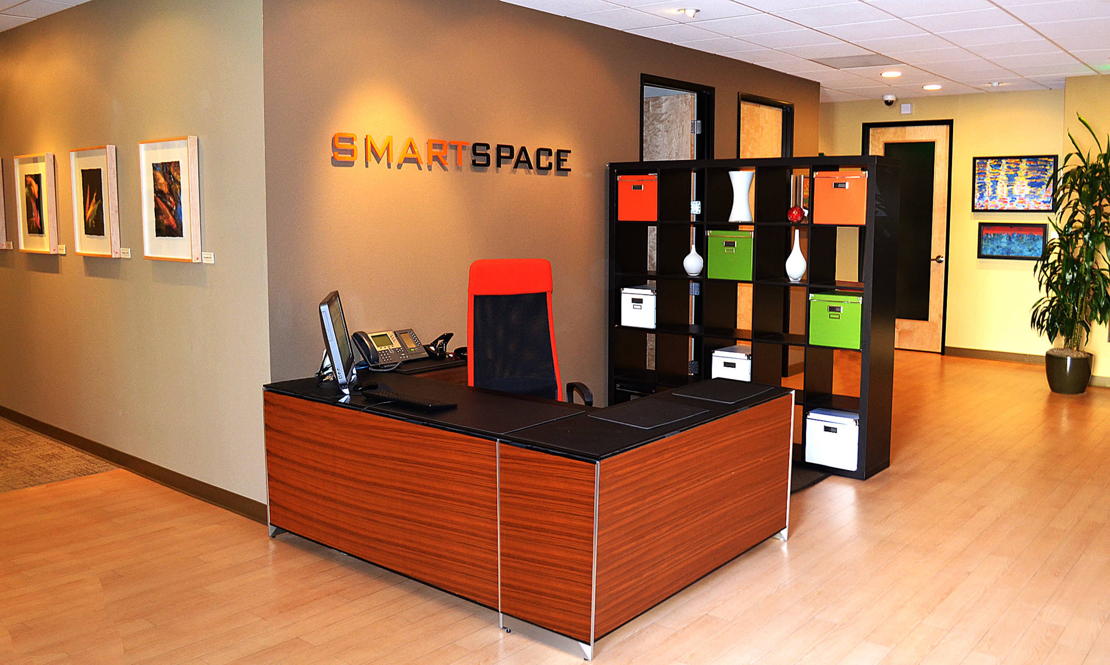 A modern office reception area featuring a desk with a red and black chair, organized storage boxes on a shelf, and framed artwork on the walls.