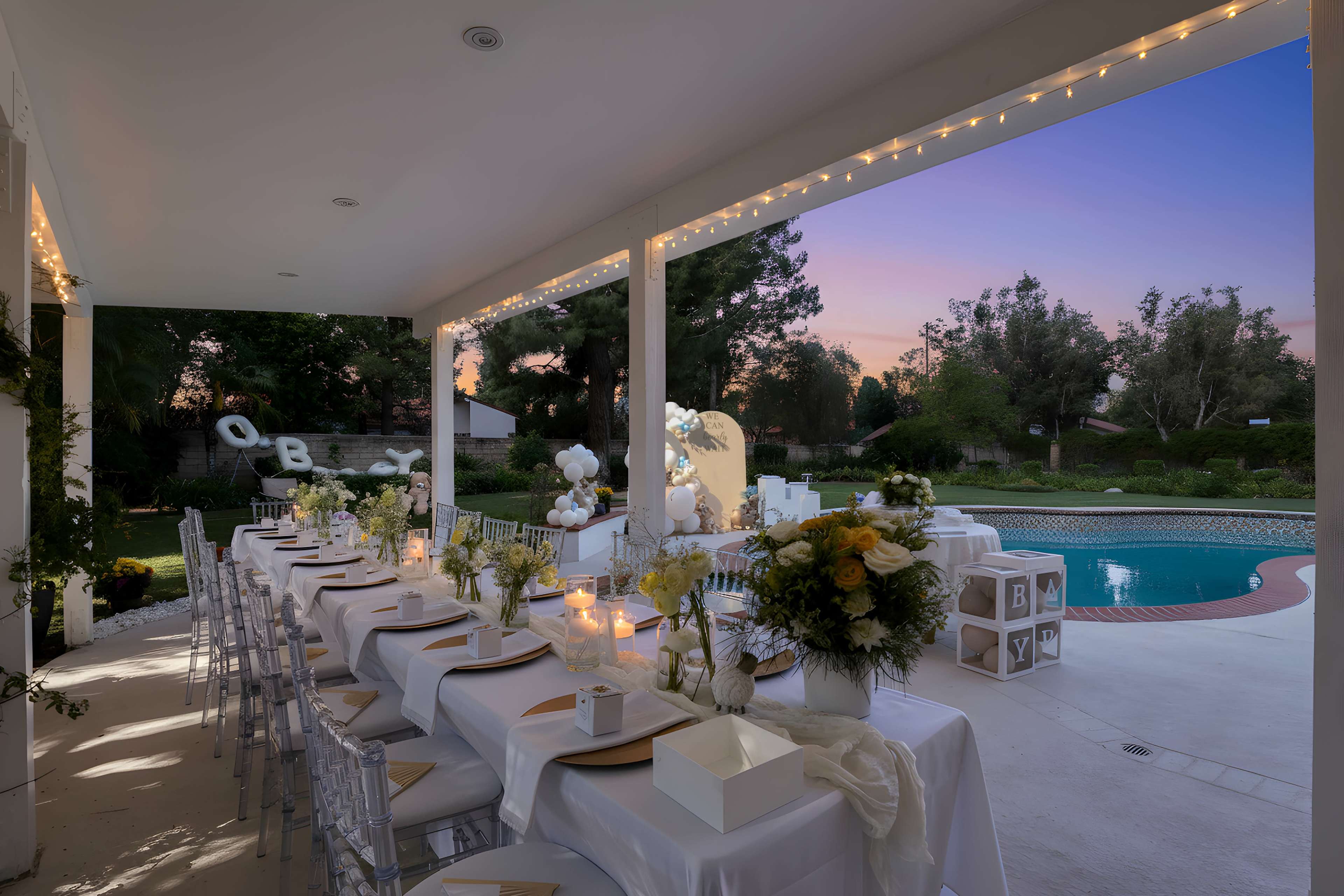 An elegantly set outdoor dining area features a long table adorned with flowers and decorations, overlooking a swimming pool and a colorful sunset in the background.