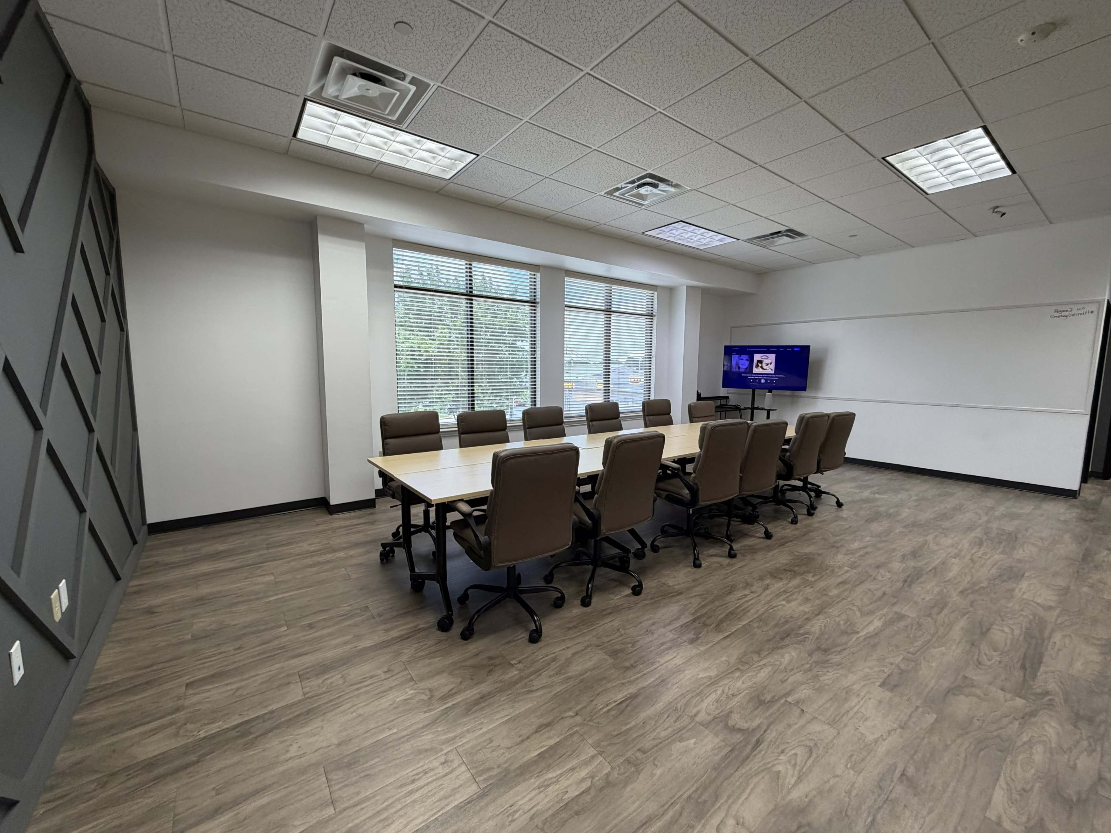 The image shows a modern conference room with a long table surrounded by ten office chairs, large windows allowing natural light, and a TV screen on one wall.