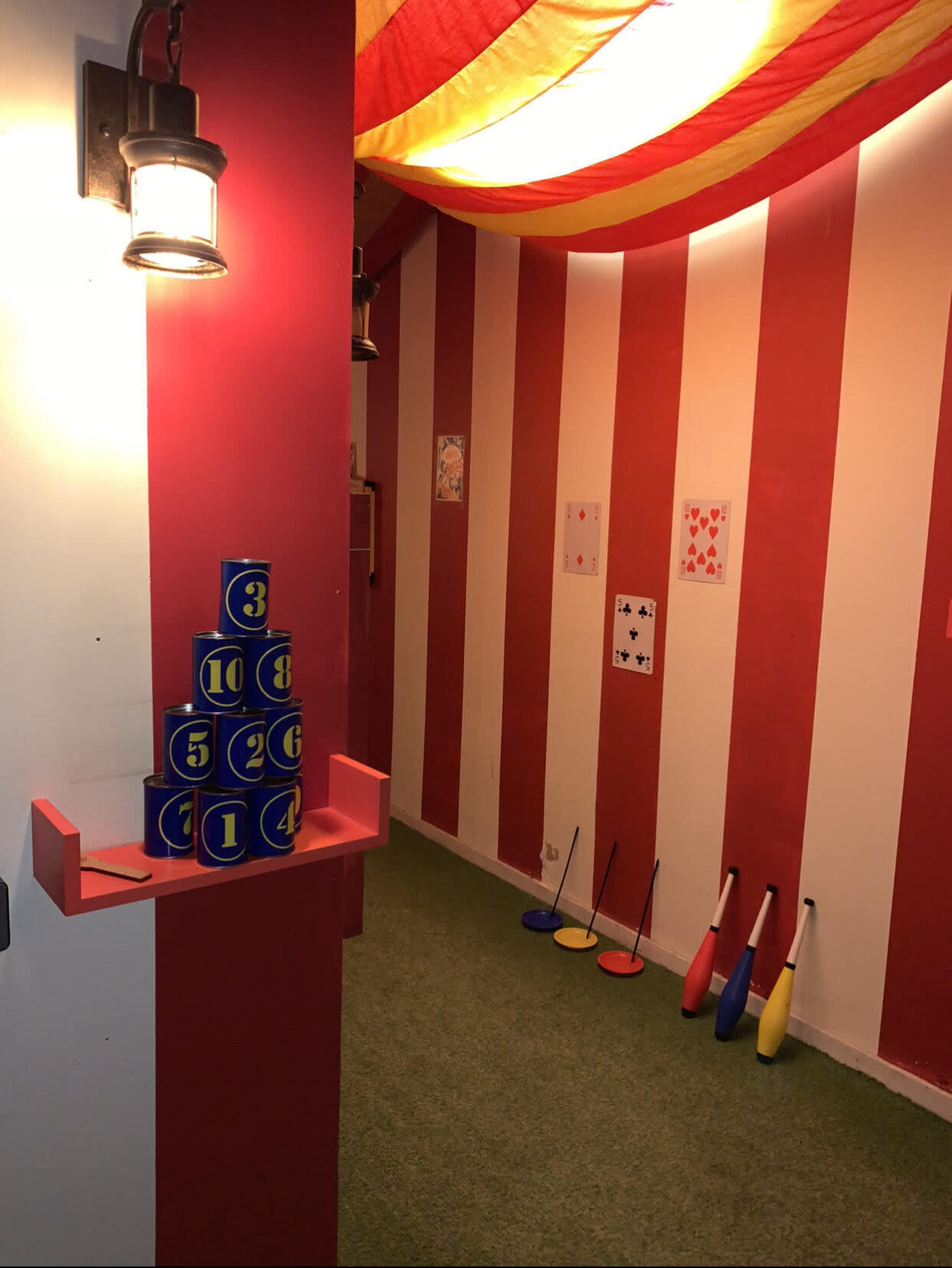 A colorful indoor carnival game area with stacked numbered blocks on the left and juggling pins arranged on the right against striped red and white walls.