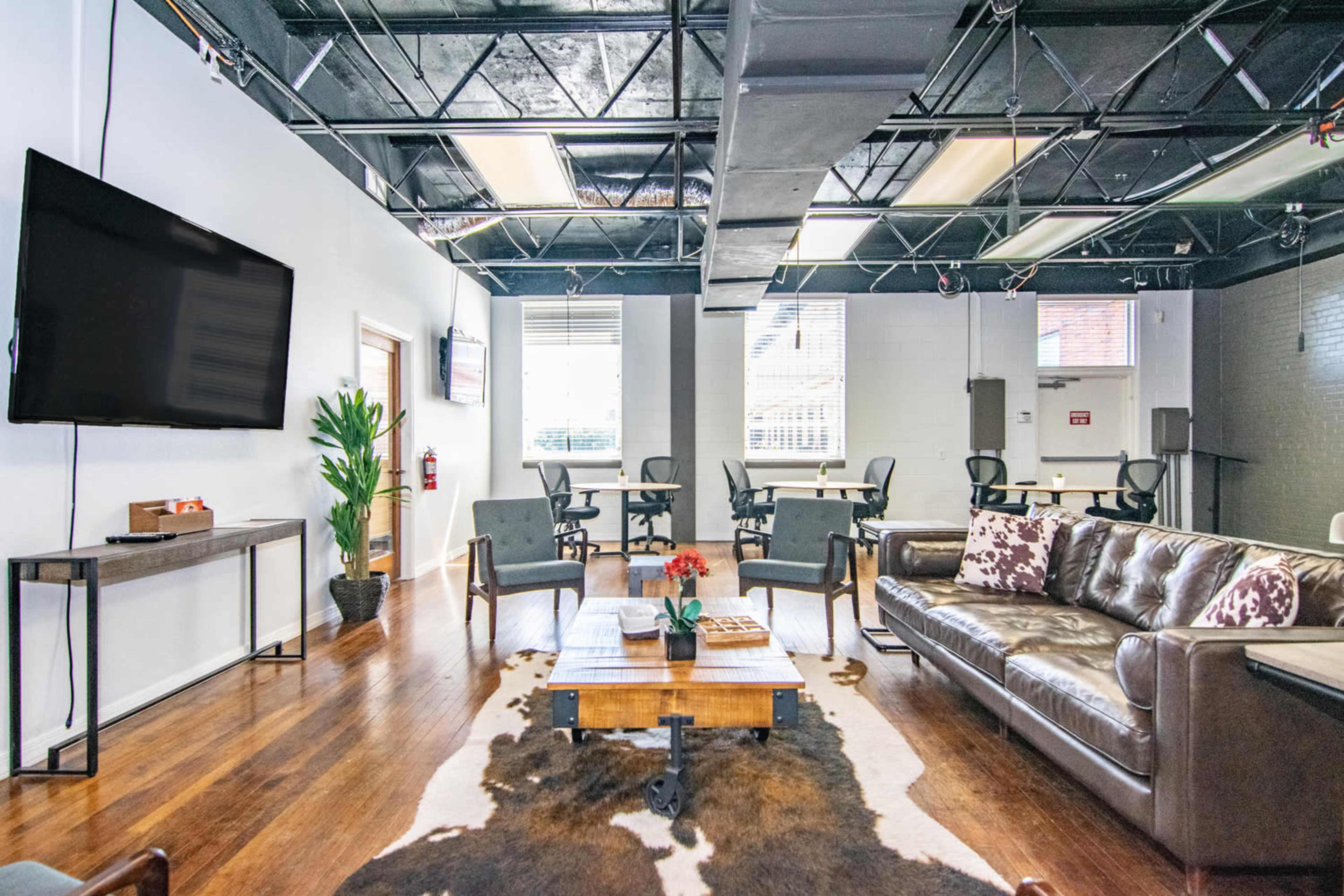 A modern office lounge features a leather sofa, a wooden coffee table, and a large television, with desks set up in the background.