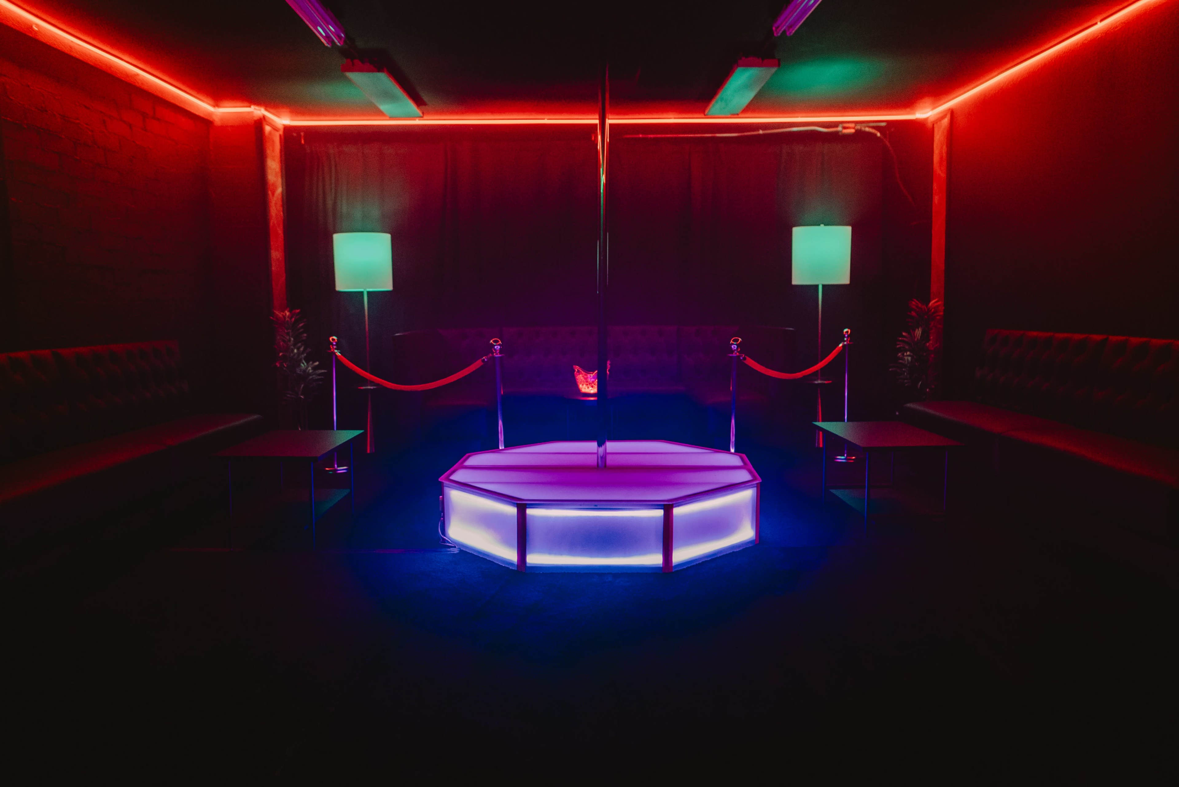 A darkened room with a illuminated octagonal stage featuring a pole, surrounded by seating areas and lit by colorful neon lights.