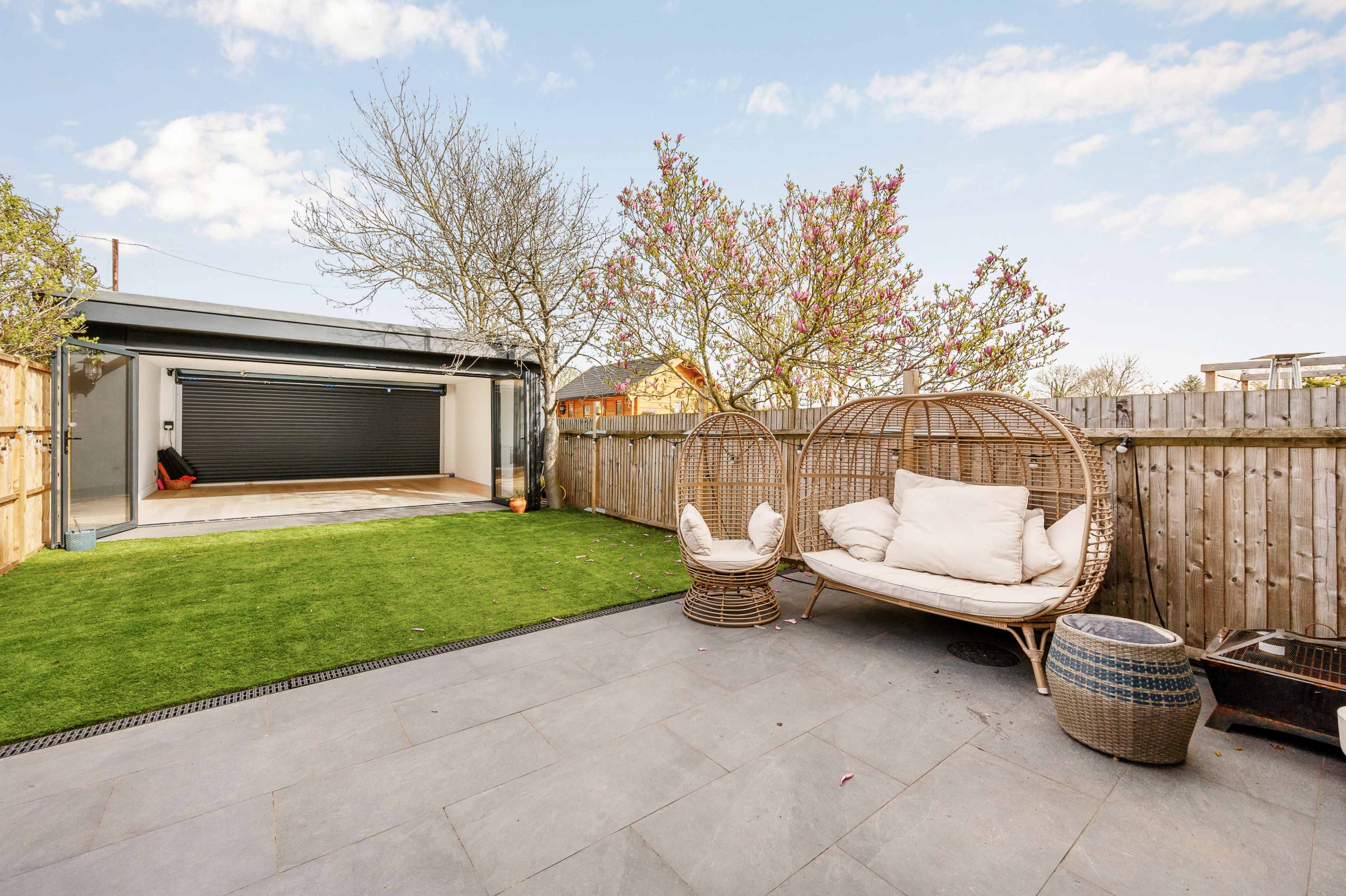 A landscaped outdoor area features two wicker chairs on a stone patio, surrounded by grass and a wooden fence, with a garage door and trees in the background.