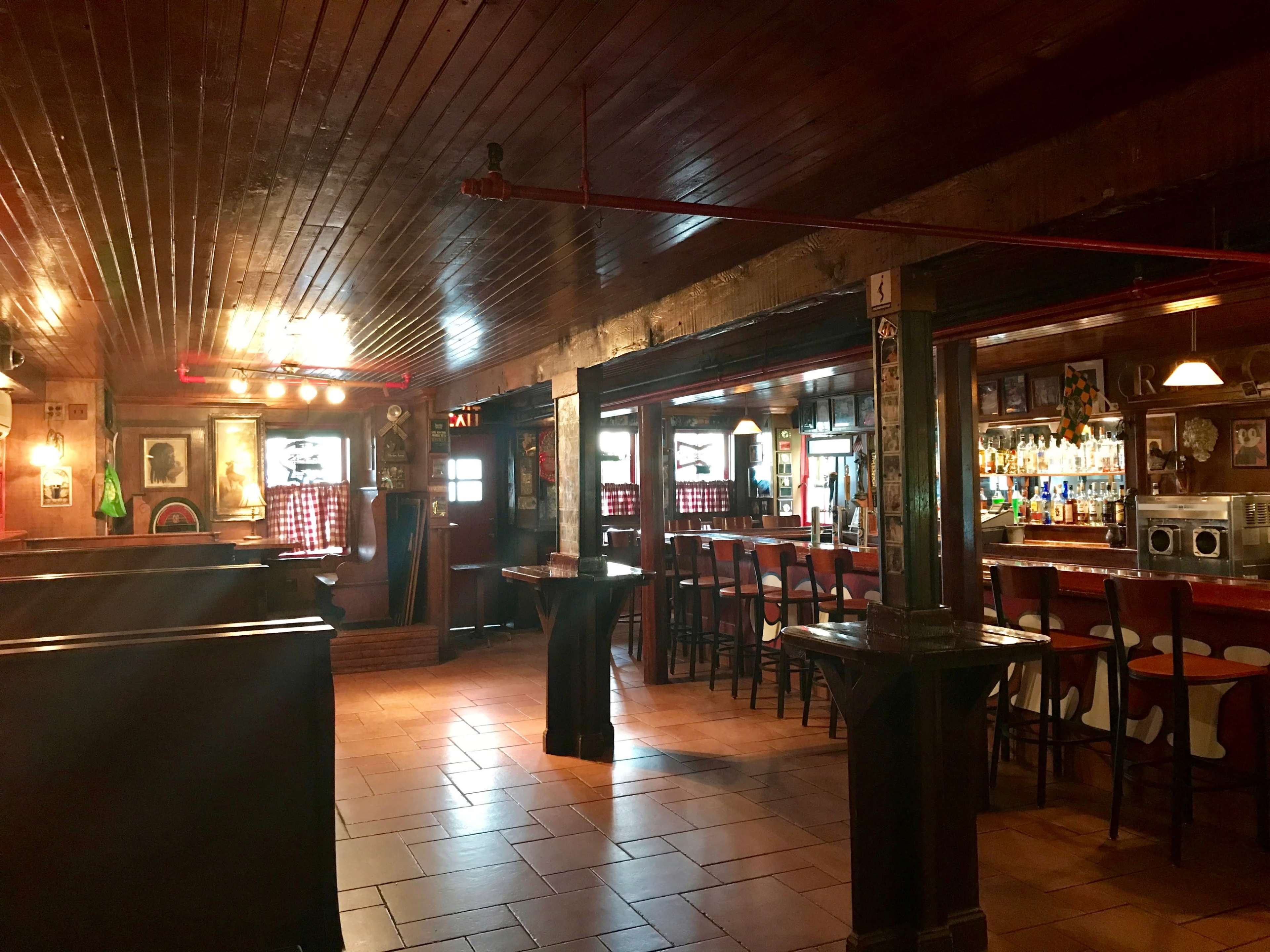The image shows the interior of a rustic bar with wooden furnishings, a long bar countertop, and empty seating areas.