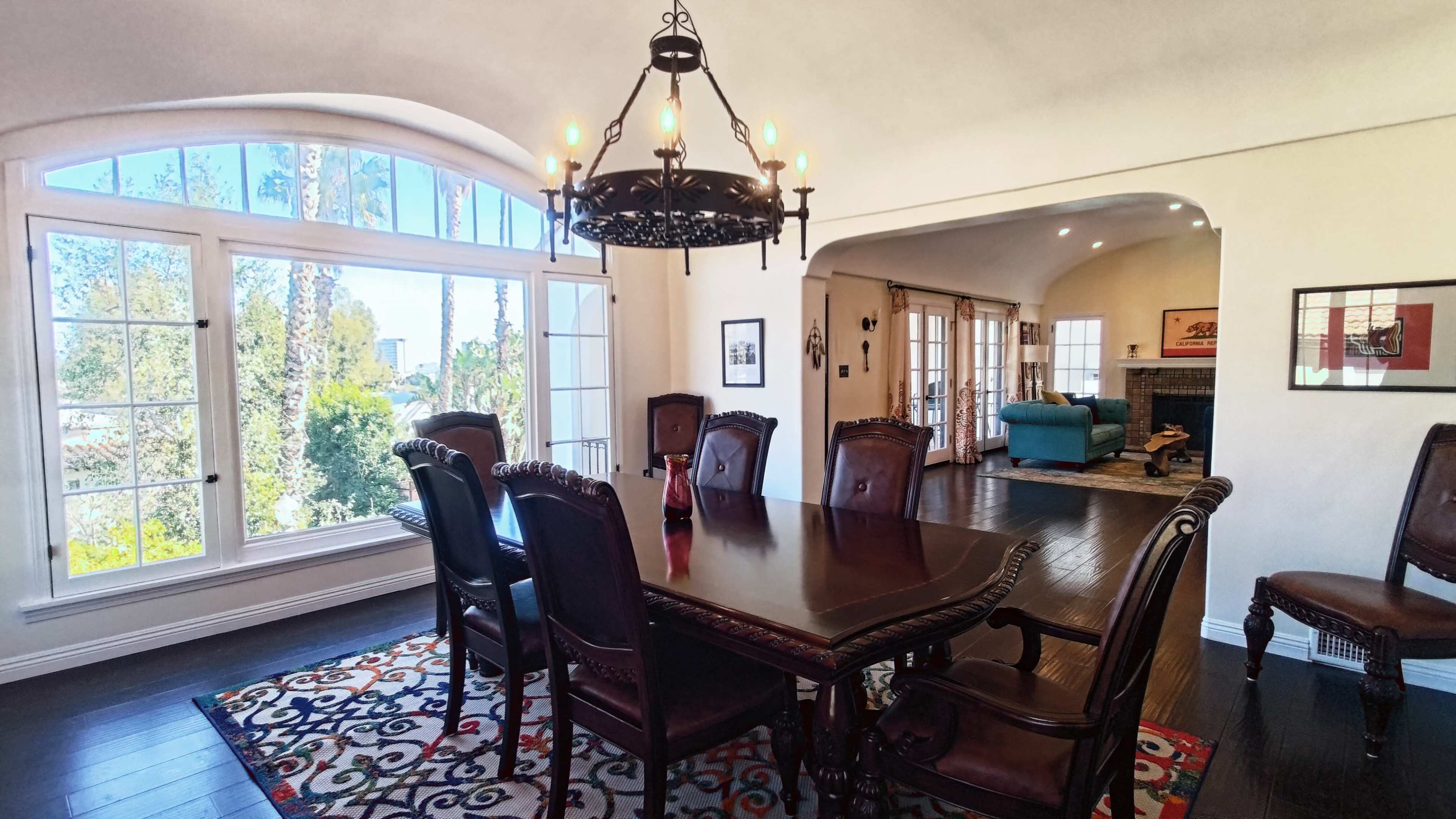 The image shows a dining room with a large wooden table surrounded by high-backed chairs, a chandelier overhead, and large windows providing natural light and a view of greenery outside.