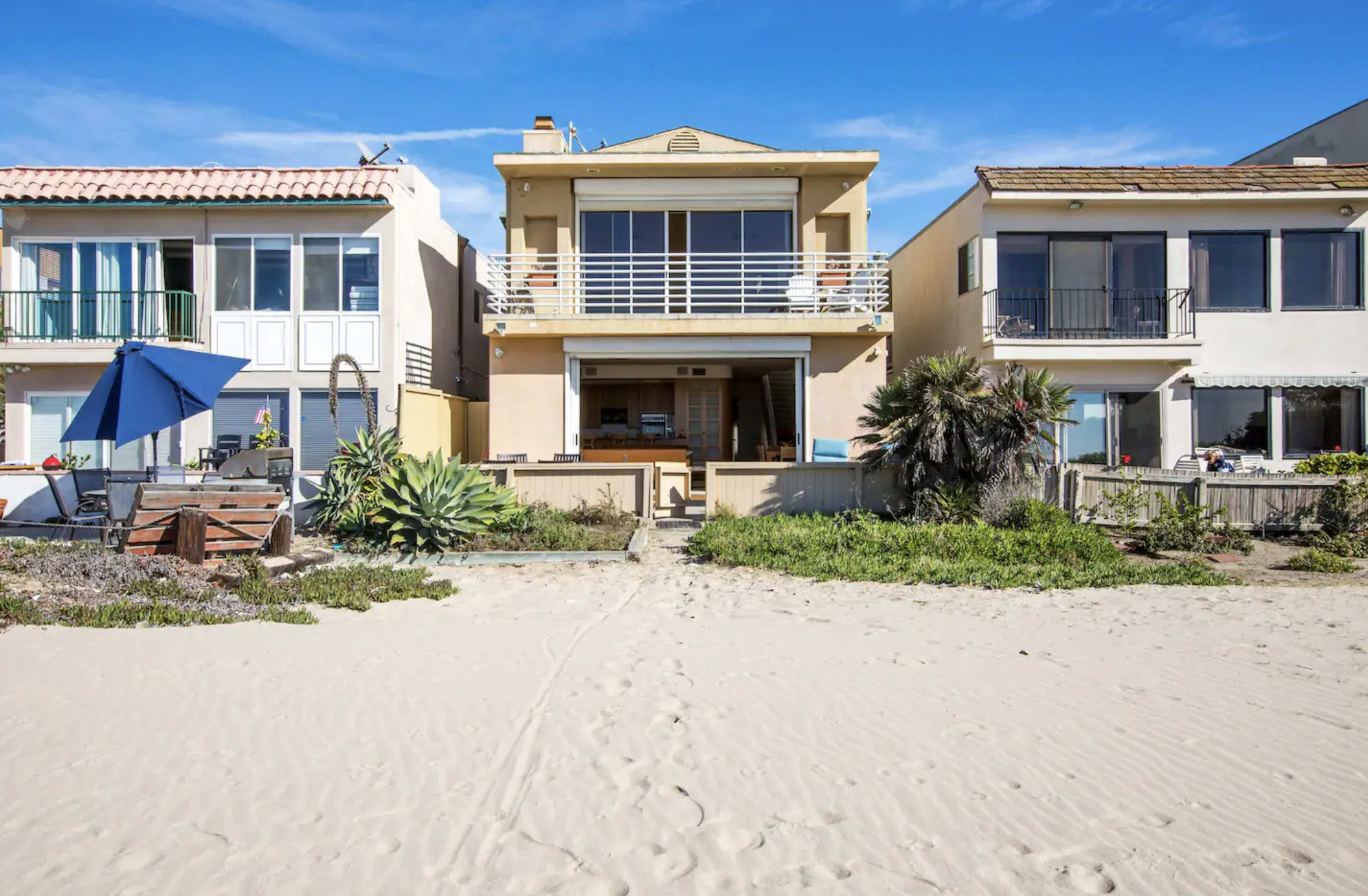 A beachfront property flanked by two neighboring houses, with sand leading up to the entrance.