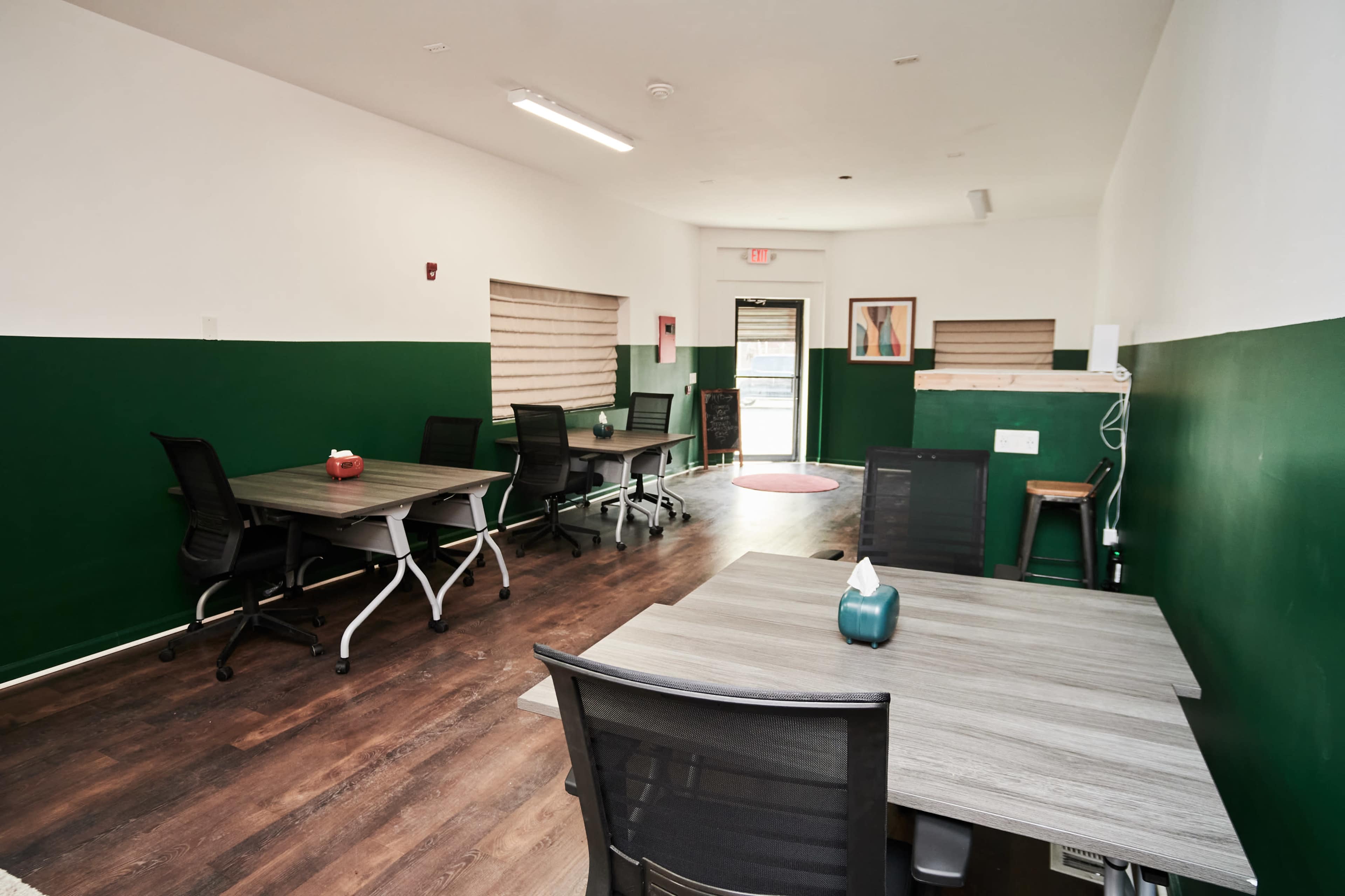 A minimalist office space with green and white walls, five desks equipped with chairs, and a small entryway.