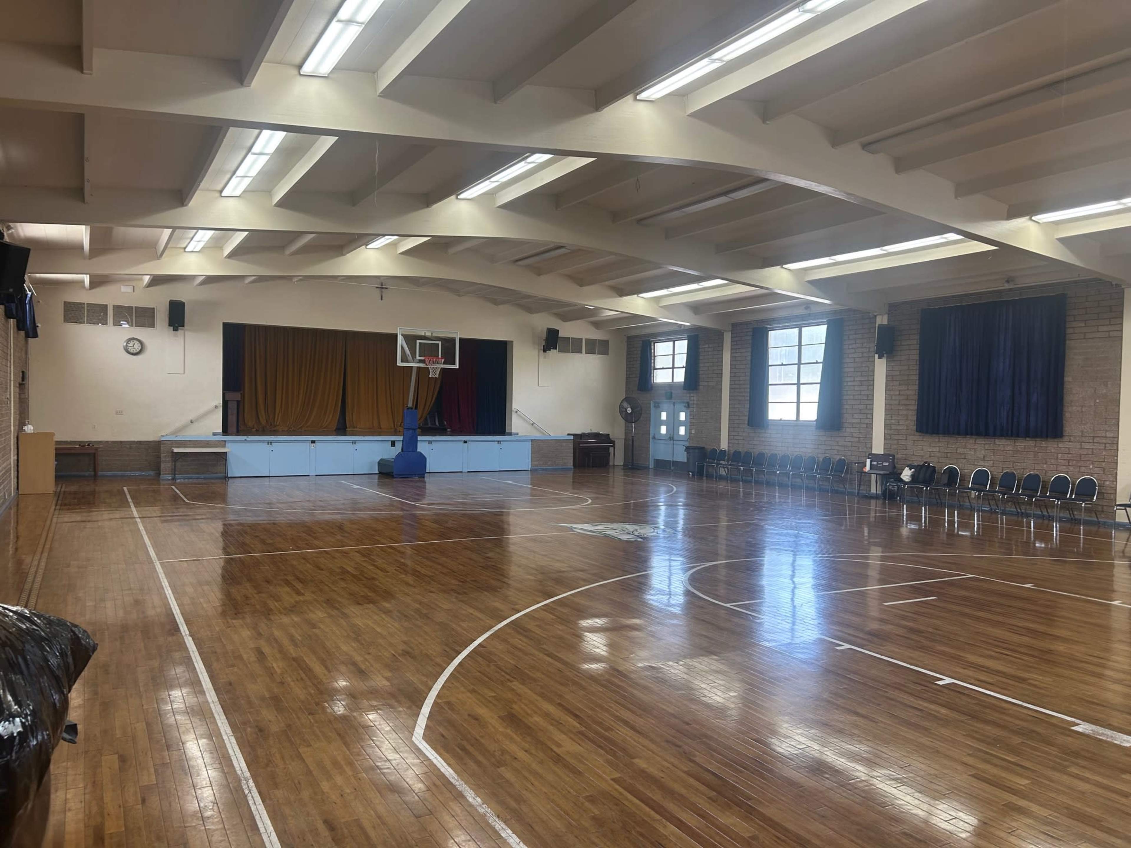 The image shows a spacious indoor basketball court with polished wooden floors, a hoop at one end, and rows of chairs lined up along the side.