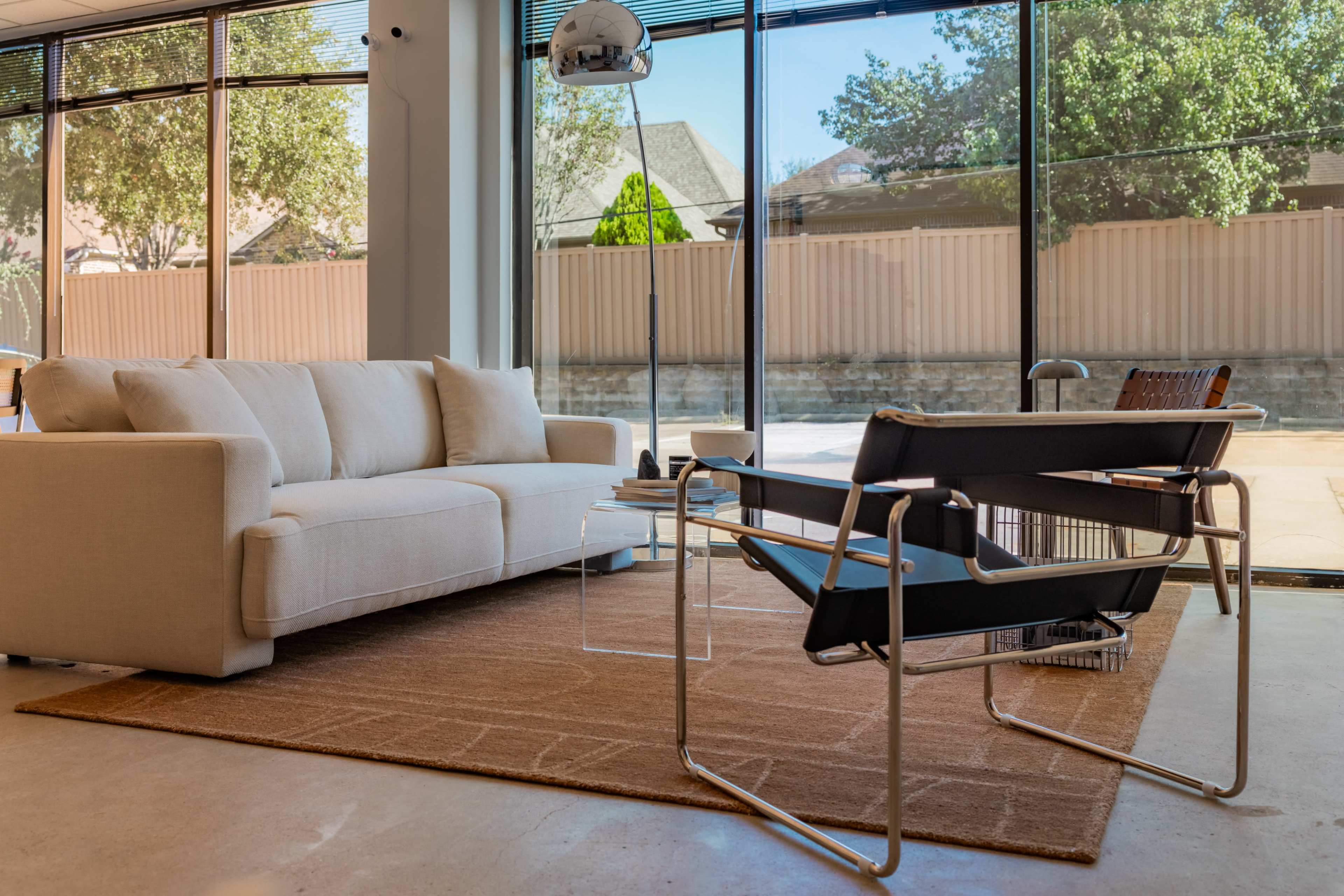 The image shows a modern living room with a beige sofa, a black lounge chair, a side table, and large windows overlooking an outdoor area.