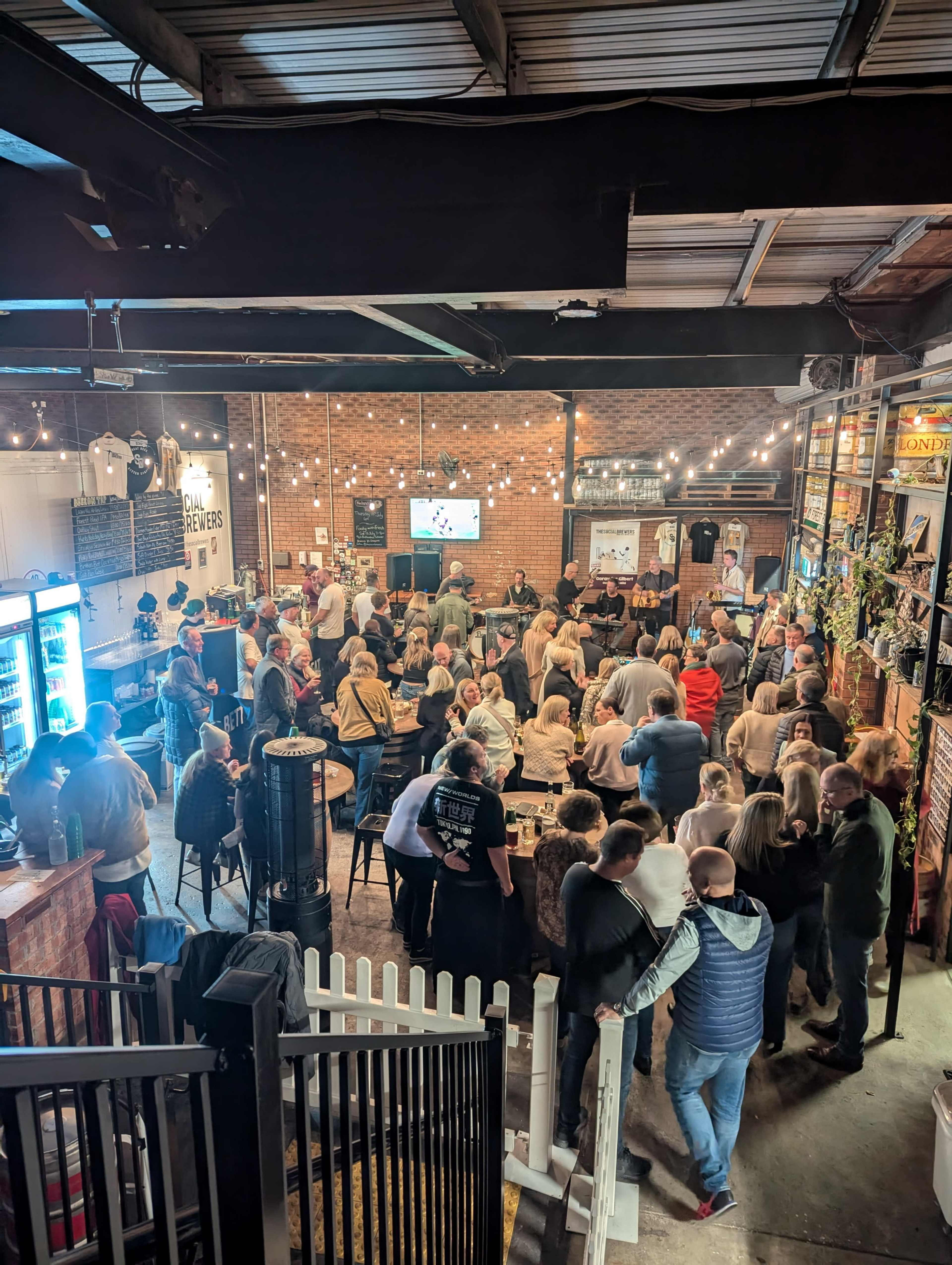 A crowded indoor venue features numerous people socializing and sitting at tables, with a large screen displaying a sports event in the background.