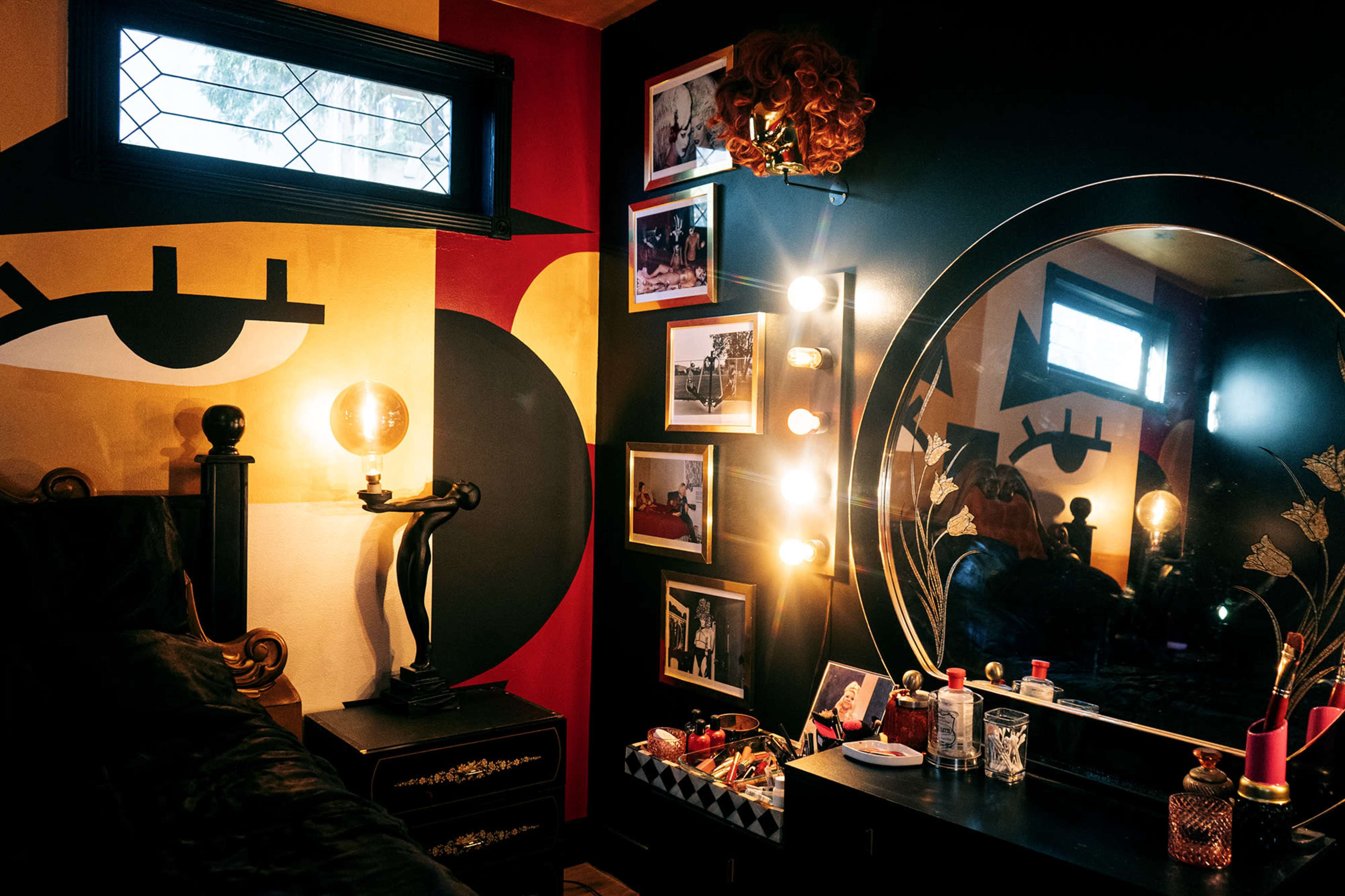 The image shows a compact bedroom featuring a bold mural on one wall, a circular mirror with light bulbs, and various framed photographs displayed alongside a bedside table.