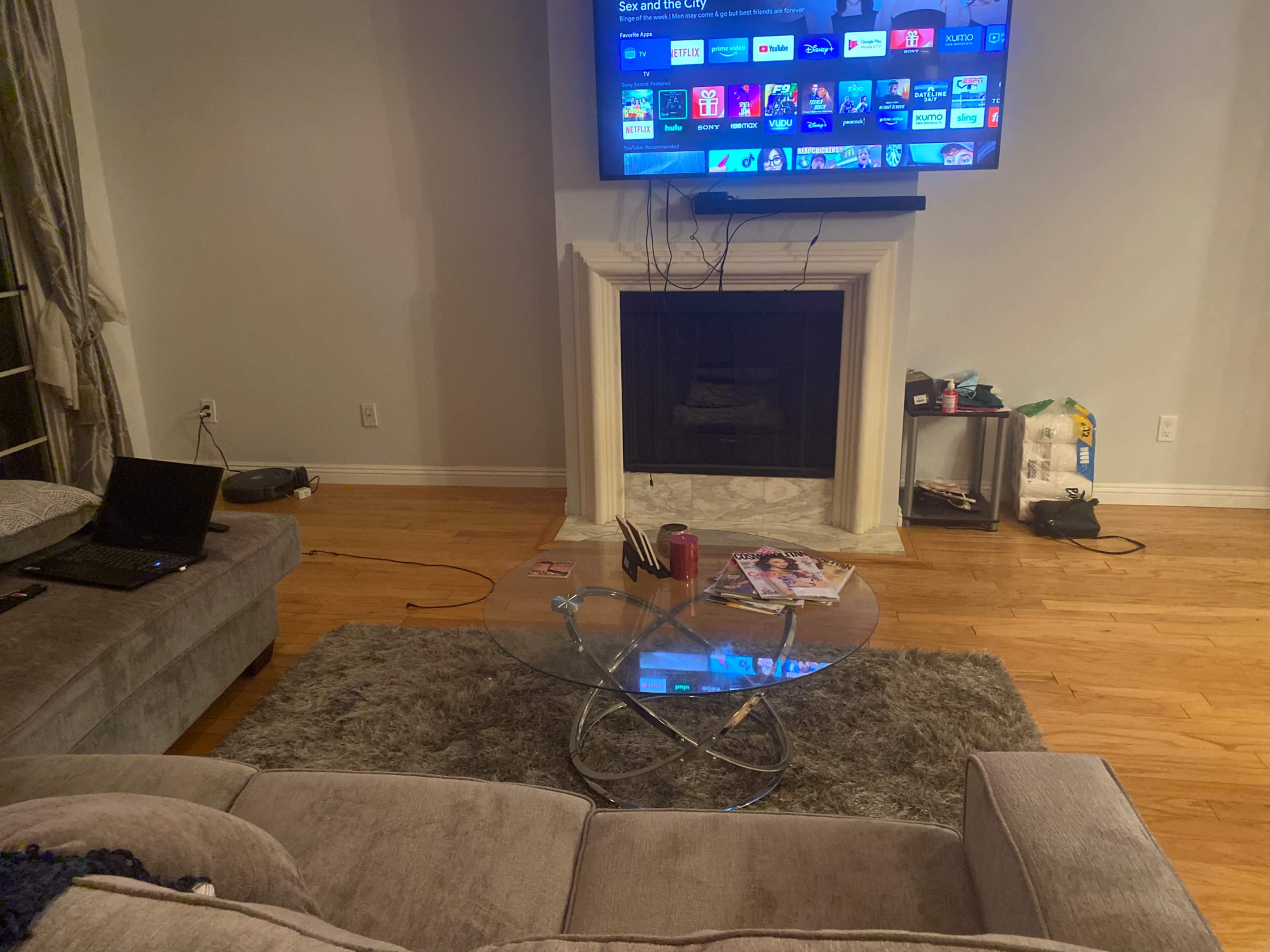 A living room features a gray sofa, a glass coffee table, a television mounted above a fireplace, and a wooden floor.