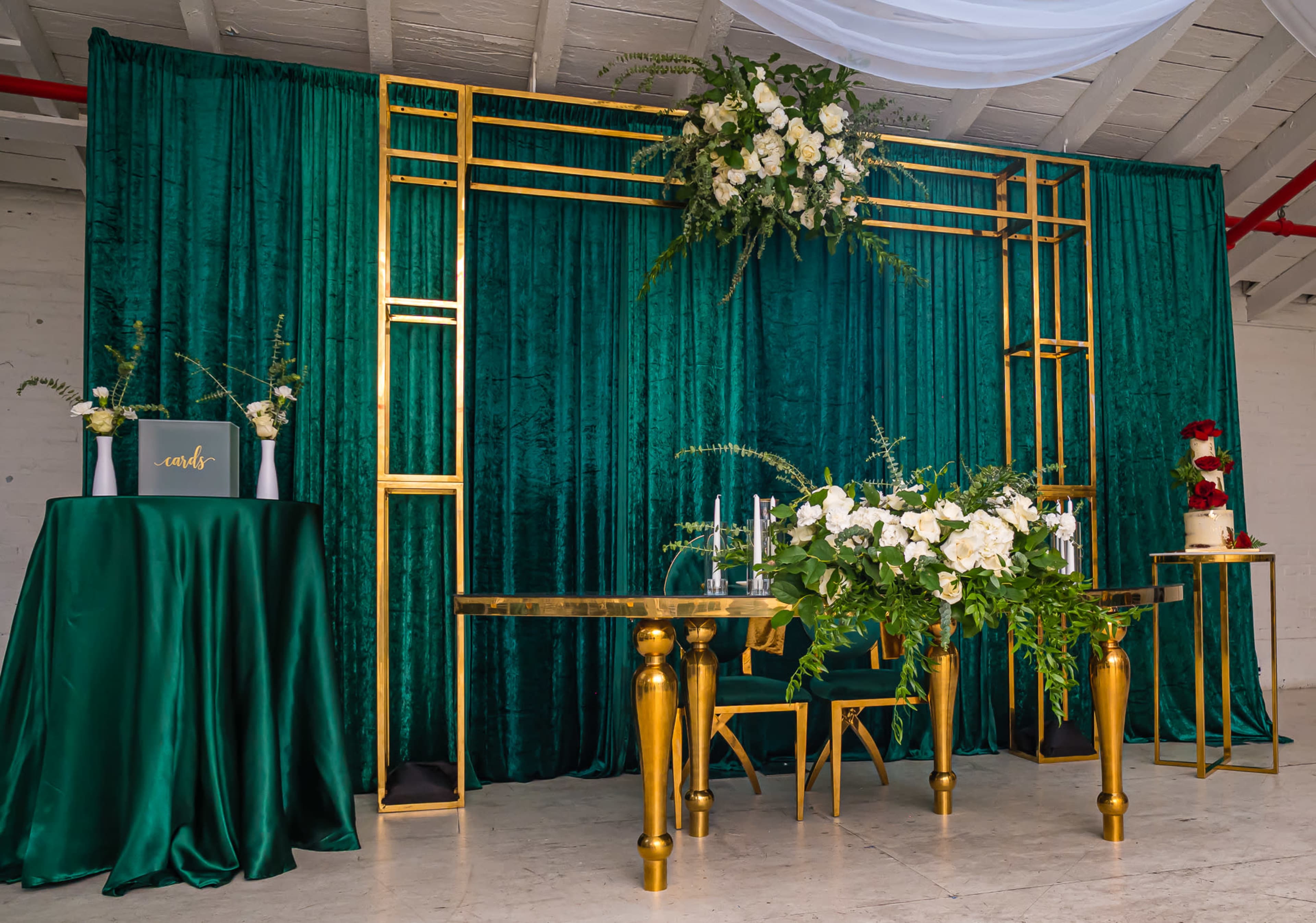 An elegant event setup with a green velvet backdrop, a gold-framed decorative structure adorned with flowers, and a table arrangement with gold accents.